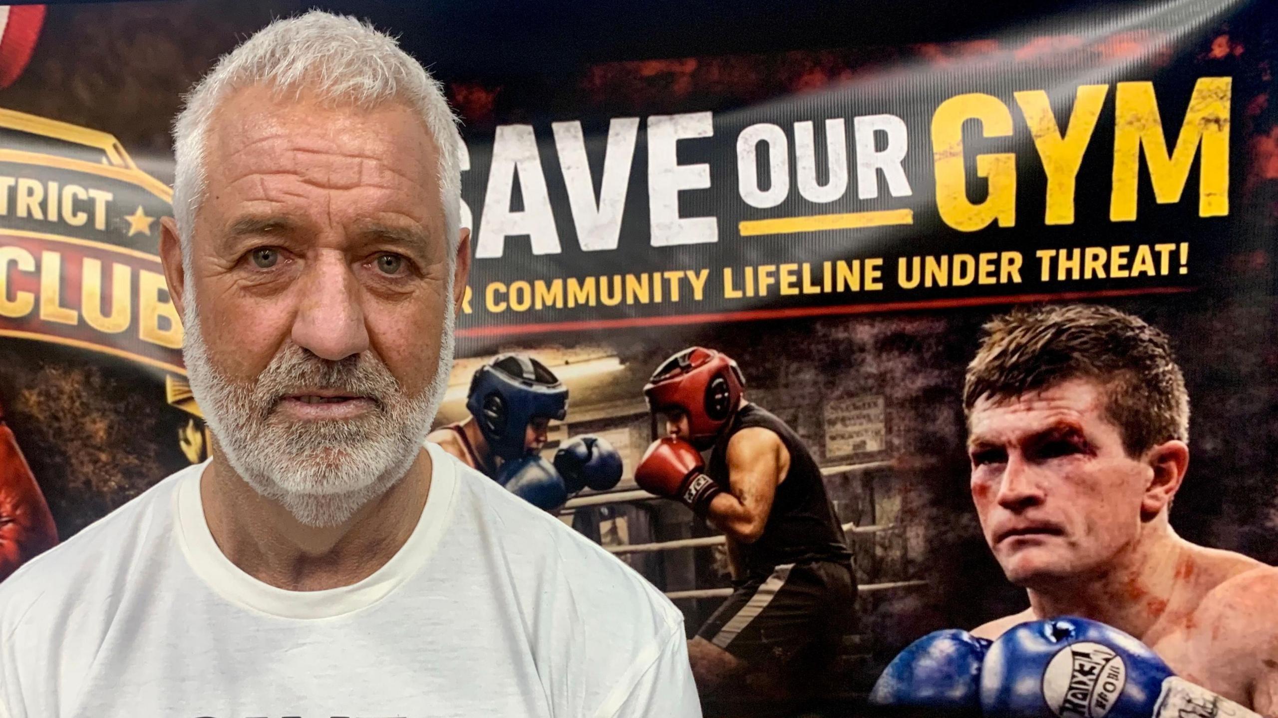 Steve Strong has white hair and a matching beard. He is wearing a white t-shirt and stands in front of a "Save Our Gym" banner which includes a photograph of two amateur boxers sparring, and a bloodied Ricky Hatton from one of his fights.