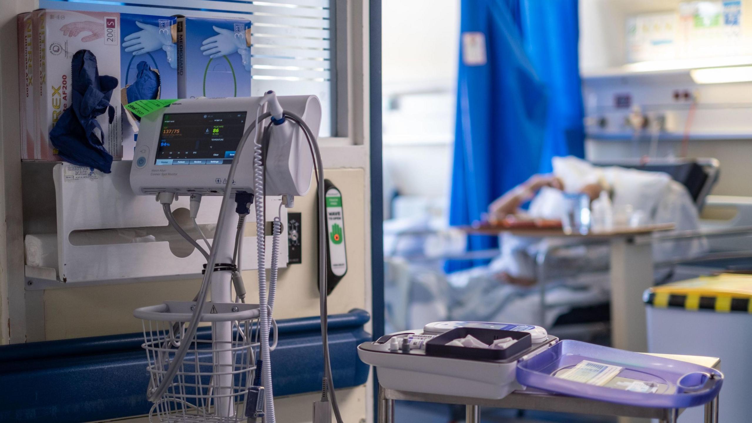 A general view of medical equipment on an NHS hospital ward. A patient can be seen out of focus in a hospital bed in the background.