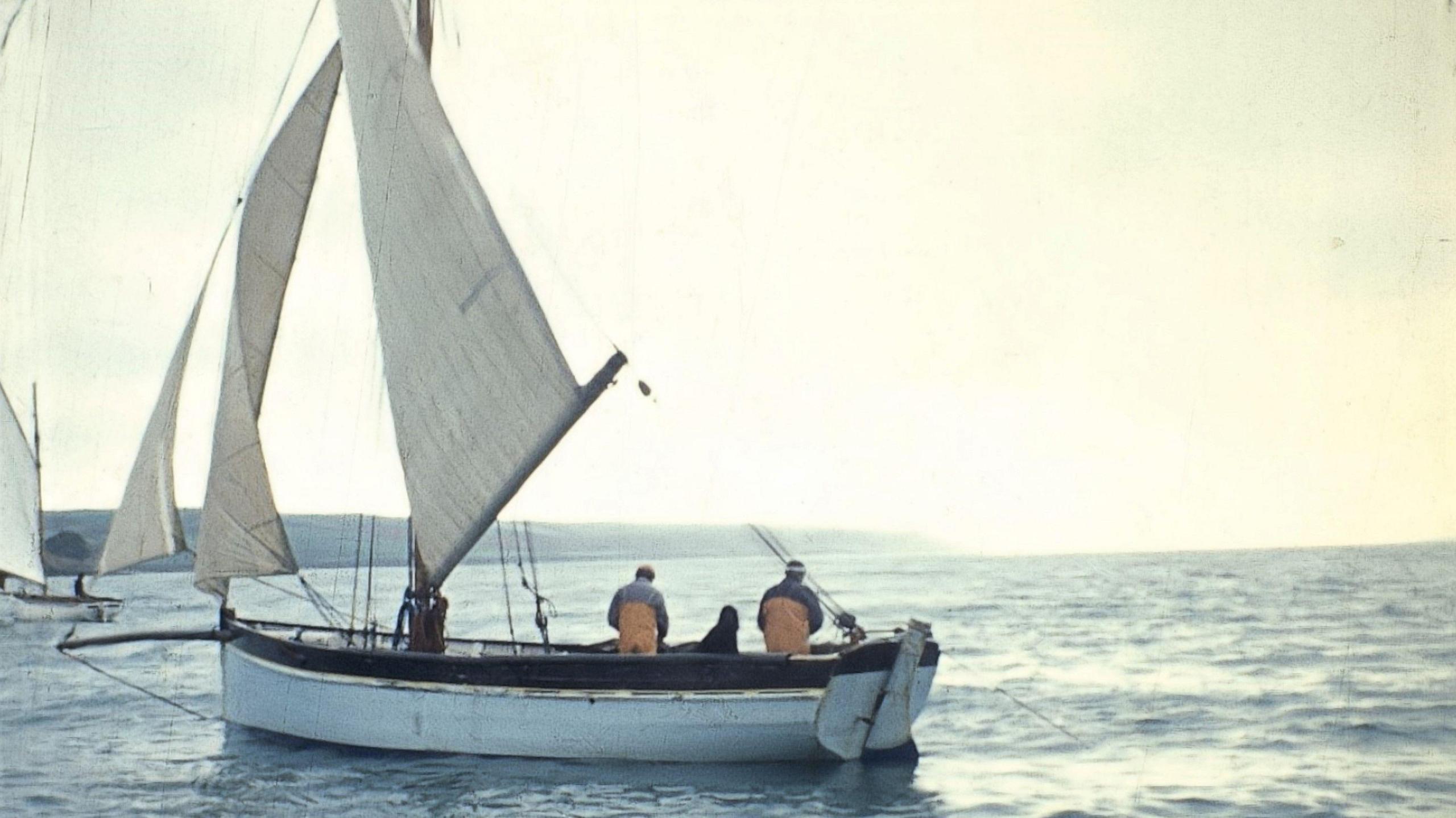 A grainy photo of a working boat under sail with two fishermen aboard. They are wearing yellow waterproof oilskins. To the left is another working boat under sail and a headland. The sea is calm and there is pale sunshine. 