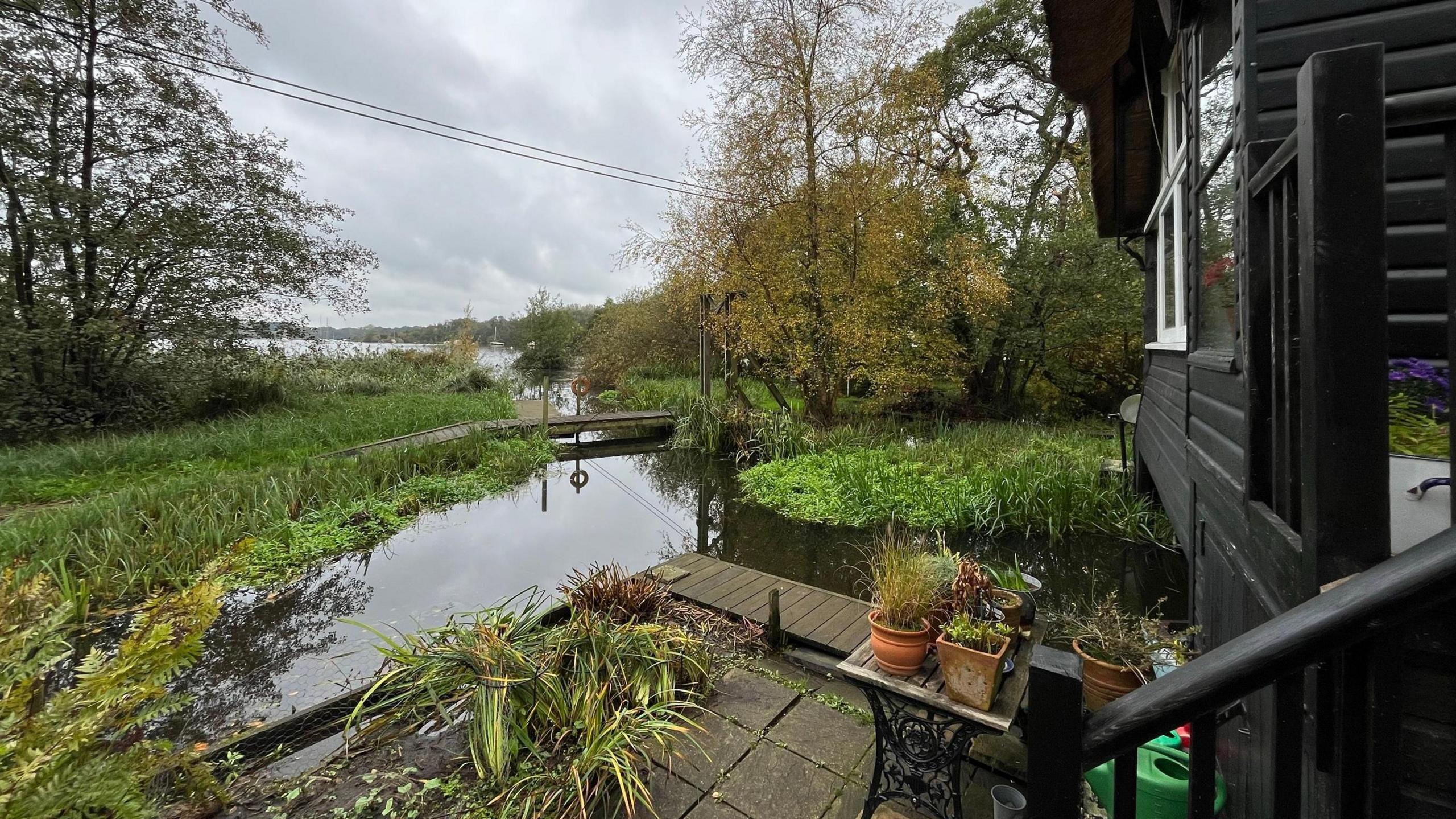 The view from a Norfolk Broads' black clad wooden building veranda showing the River Bure coming up in front of the house and bearing to its right to go under the house. Trees can be seen further off on either side of the river, which then can be seen joining a larger mass of water