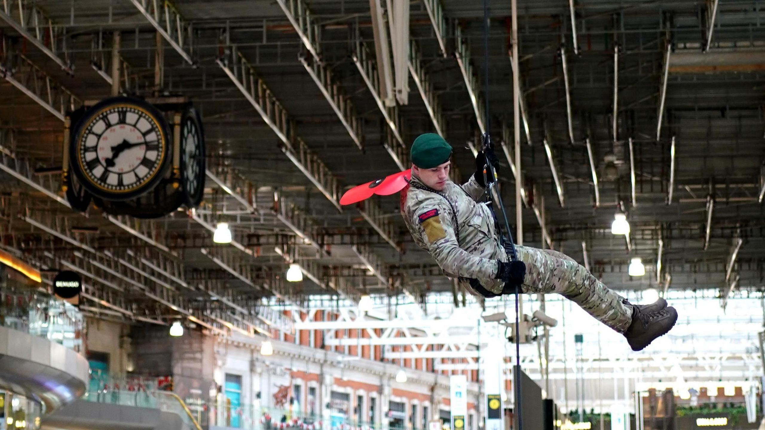 A member of the Royal Marines abseils at the launch of the Royal British Legion's London Poppy Day, at Waterloo Station in London. The image shoes a Royal Marine with a green beret and a large model of a red poppy attached to his back. He is holding a rope and looking down.