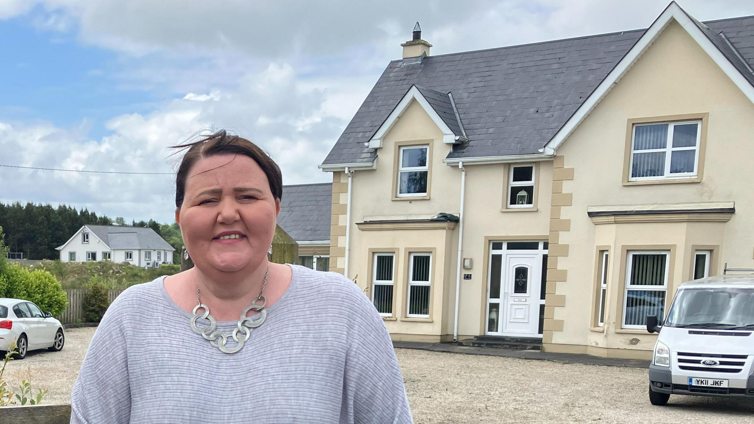 Joy Beard standing outside her home before demolition started. Her dark hair is in a ponytail and she is wearing a grey jumper and a silver necklace. The house is a large, detached, cream home with two bay windows on the bottom.