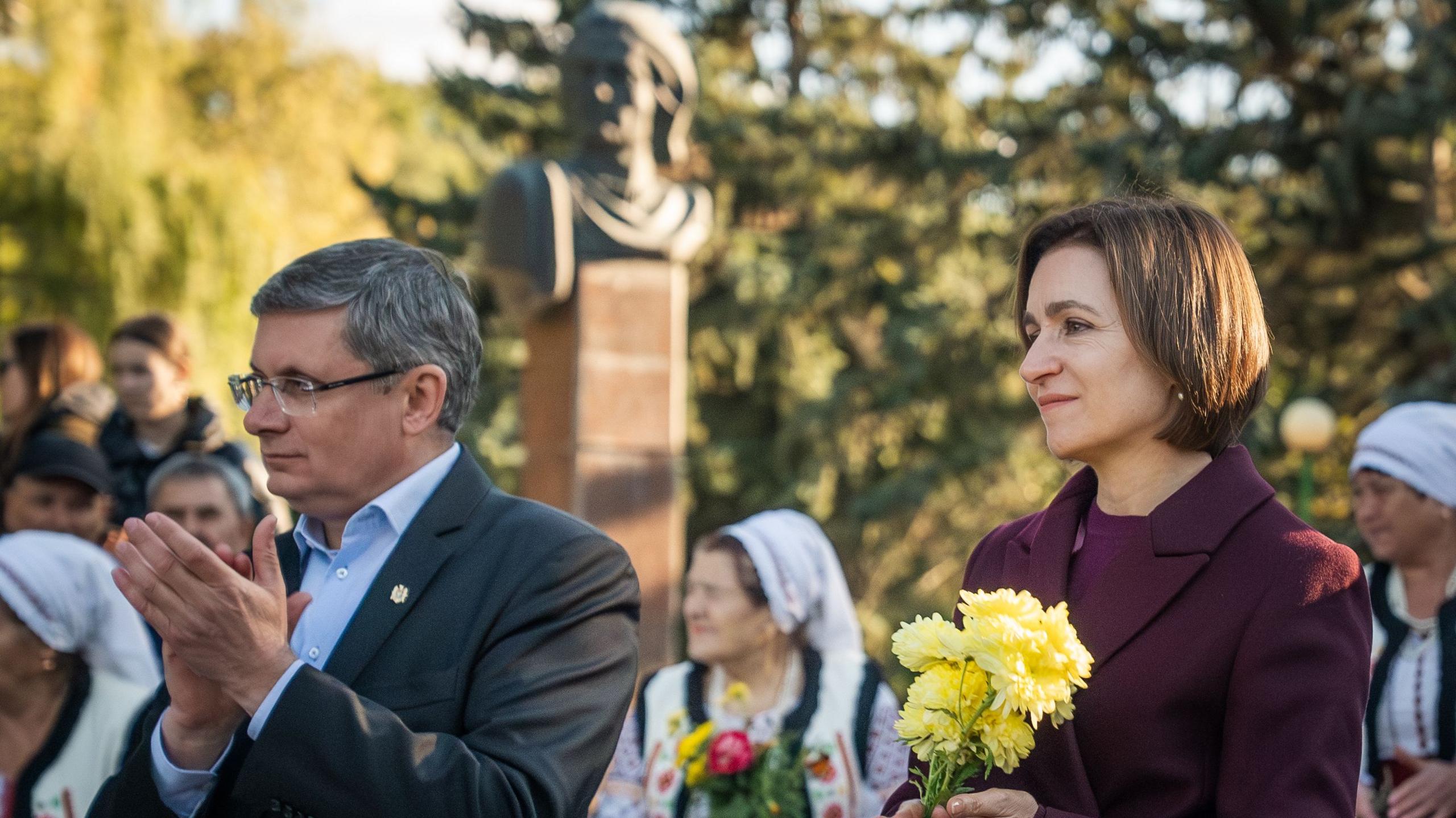 President Maia Sandu holding a small bunch of flowers during a campaign visit. She is wearing a purple suit jacket. A man standing on the left is applauding. There are people dressed in traditional clothing in the background.