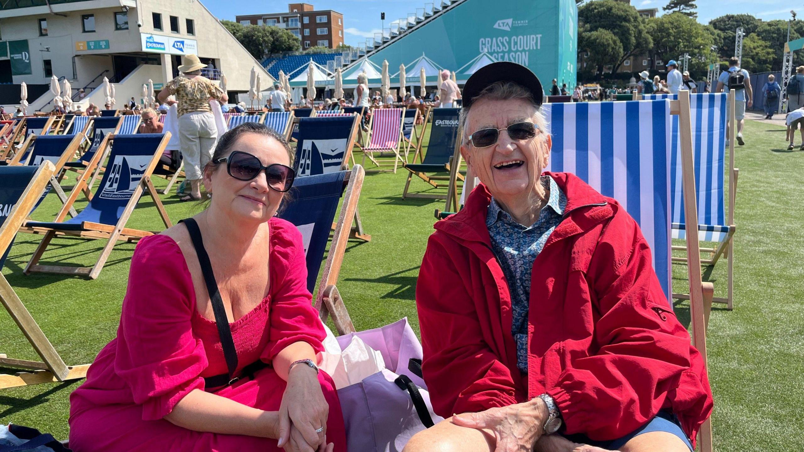 George and Julie Bell, both dressed in red and wearing sunglasses, sit in deck chairs outside Devonshire Park in Eastbourne and smile into the camera.