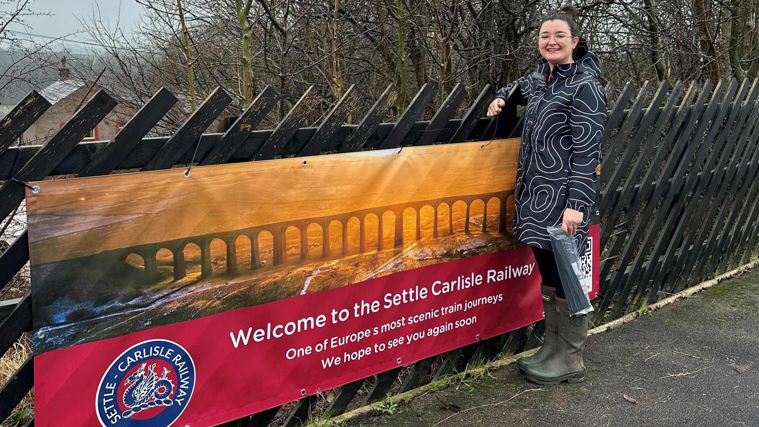 A young woman stands next to a banner attached to a station fence. The banner depicts the Ribblehead Viaduct at sunset and welcomes passengers to one of Europe's most scenic train journeys. The woman has dark hair tied back and wears glasses, a black and white raincoat, black leggings and green wellies. She is holding a bag of cable ties and smiling.