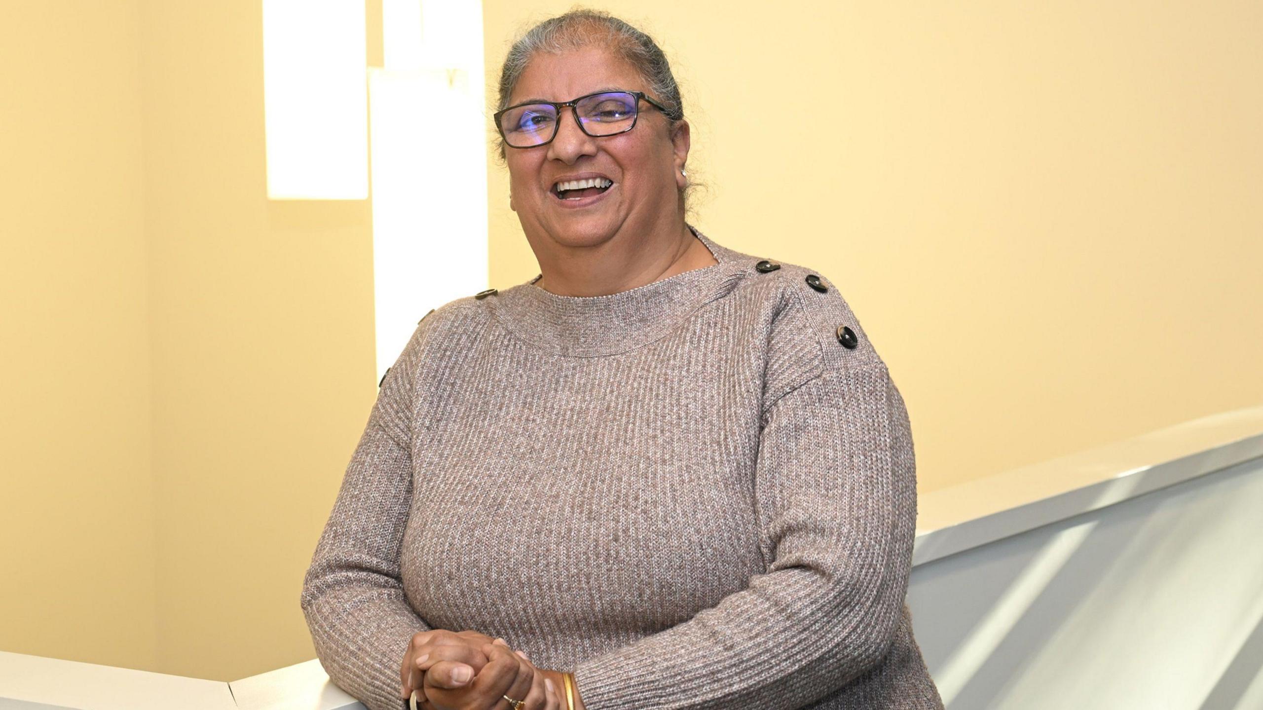 Mr Kaur stands against a pale yellow backdrop, smiling. She has grey hair worn up, and wears black-framed glasses and a beige knitted jumper with tortoiseshell buttons along the shoulders.