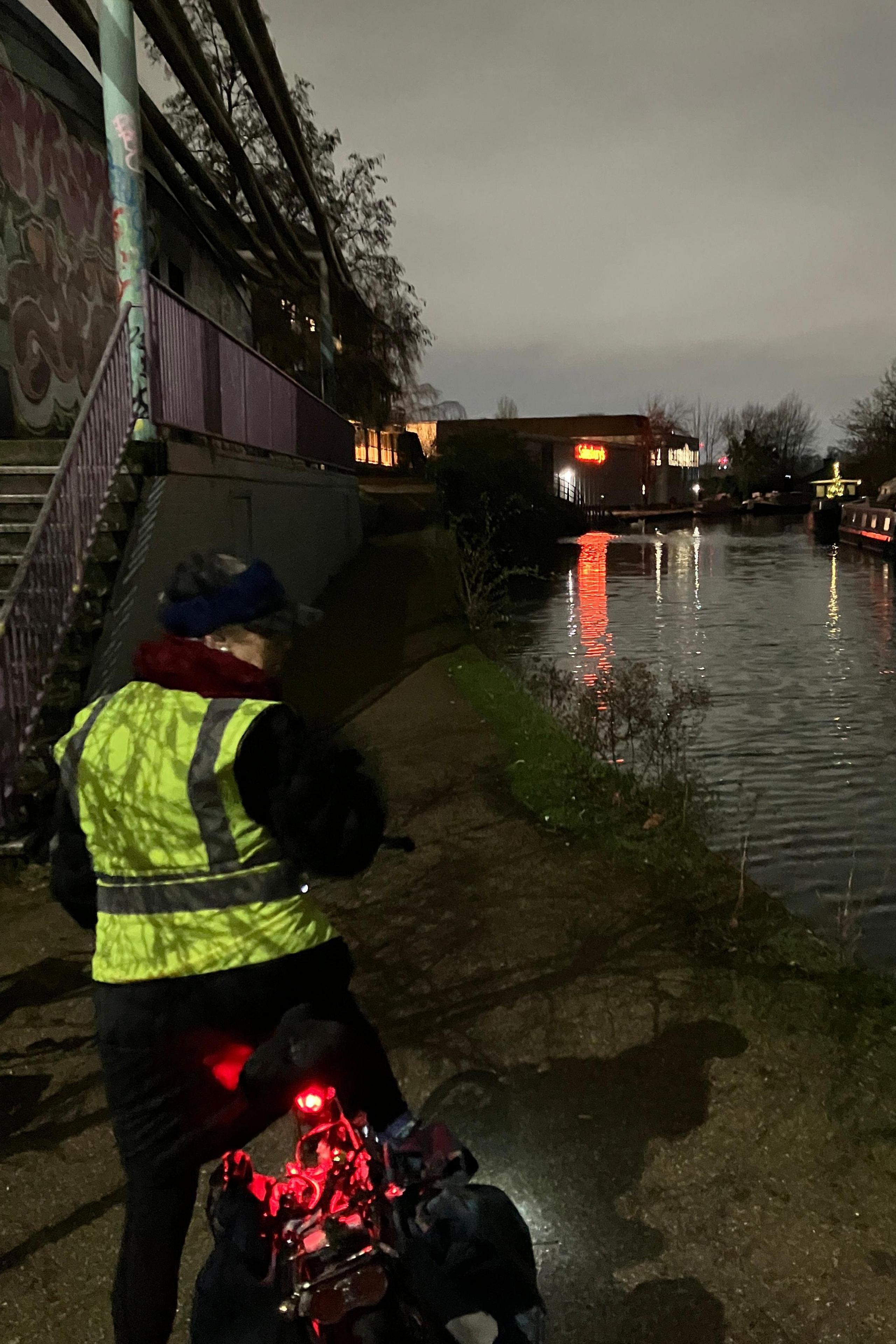 Back view of female cyclist paused at the edge of a canal. She wears yellow hi-vis jacket and her red rear cycle light glows in the dusk.