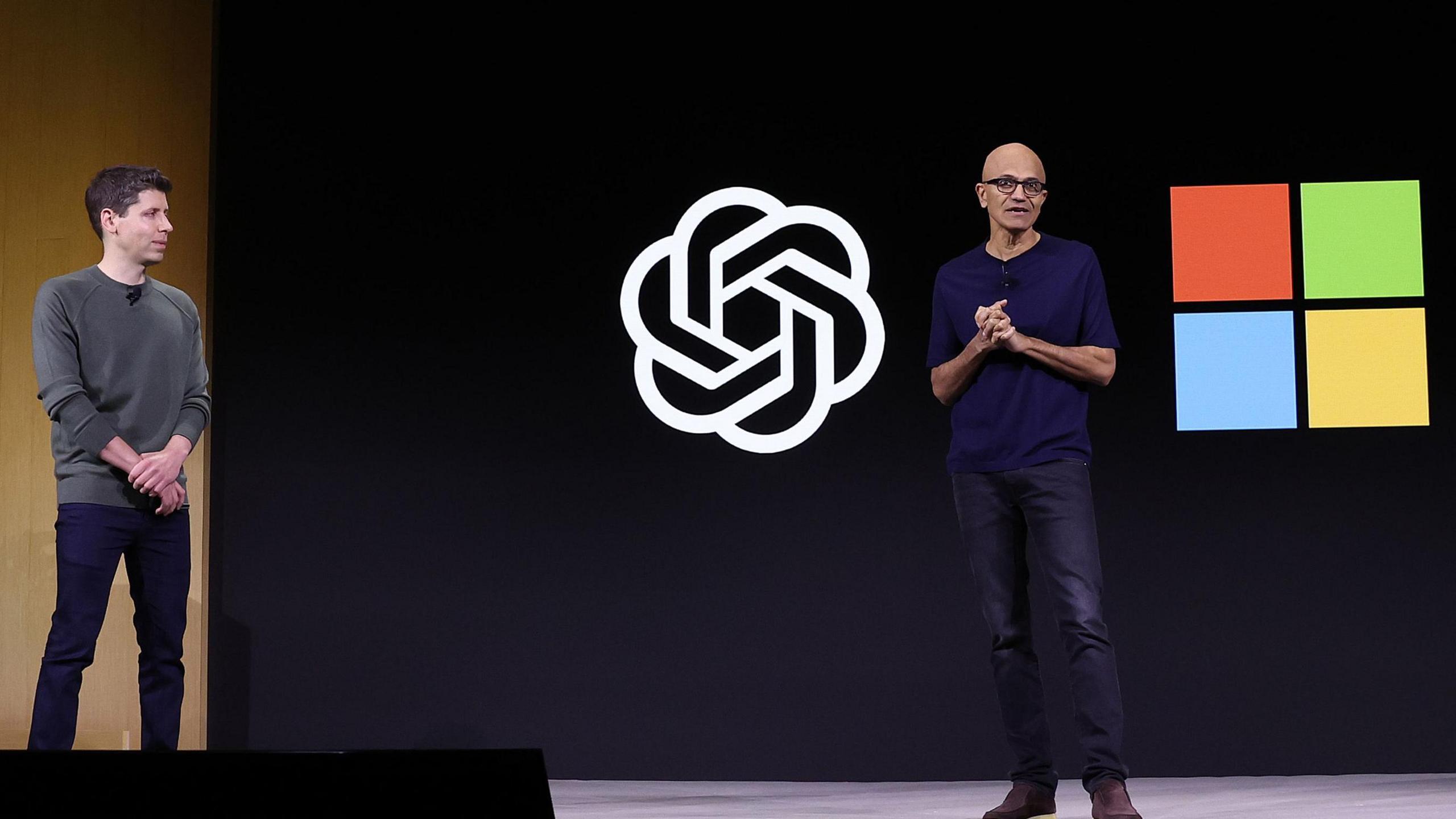 Sam Altman and Satya Nadella standing on a stage in front of logos of their two companies. Both are wearing jumpers and jeans.