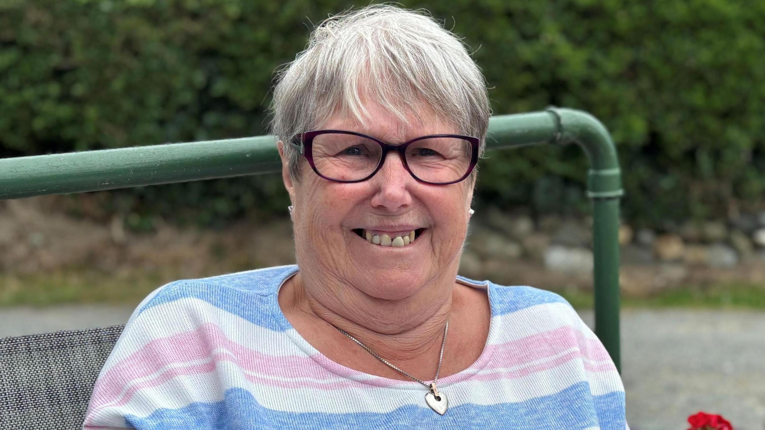 Close up image of Helen smiling looking at the camera. She is sat on a bench outside. Helen is wearing a long sleeve top with white, blue and pink stripes. She has short grey hair and wears purple glasses. It is a head and shoulders shot of her.