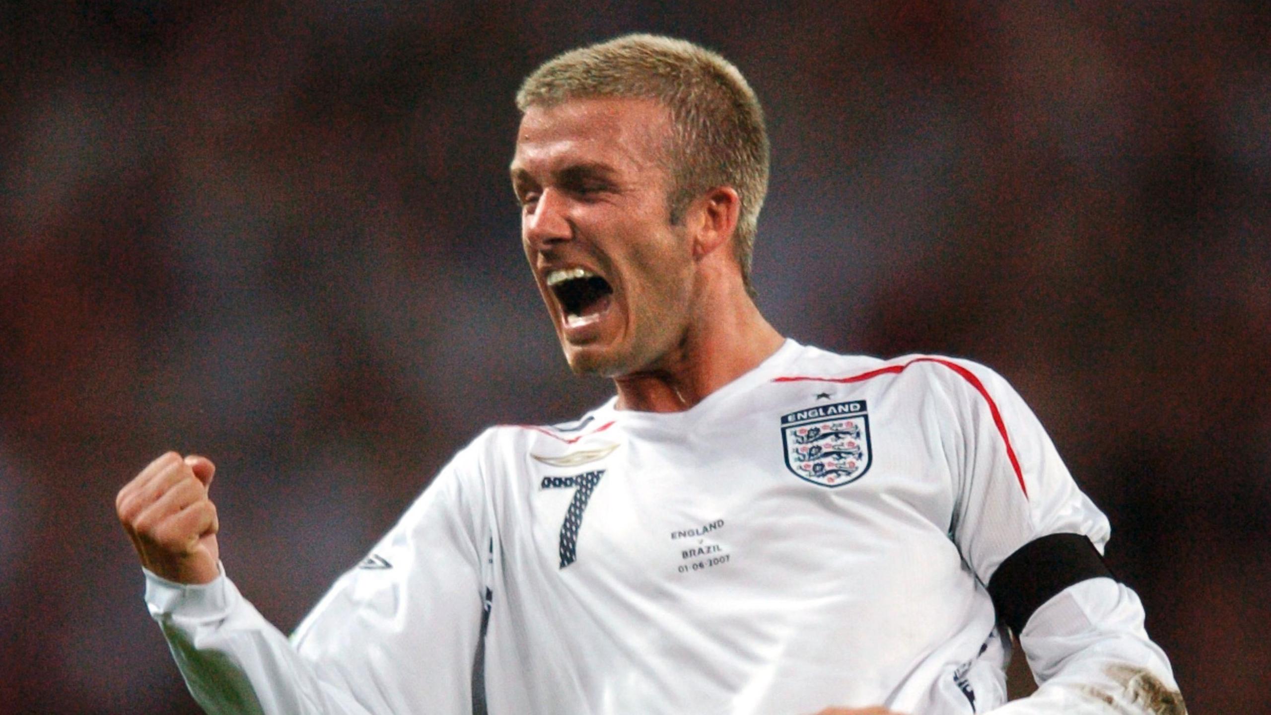 Sir David Beckham wearing a white, long-sleeved England football top during a match. He is cheering and fist pumping the air.
