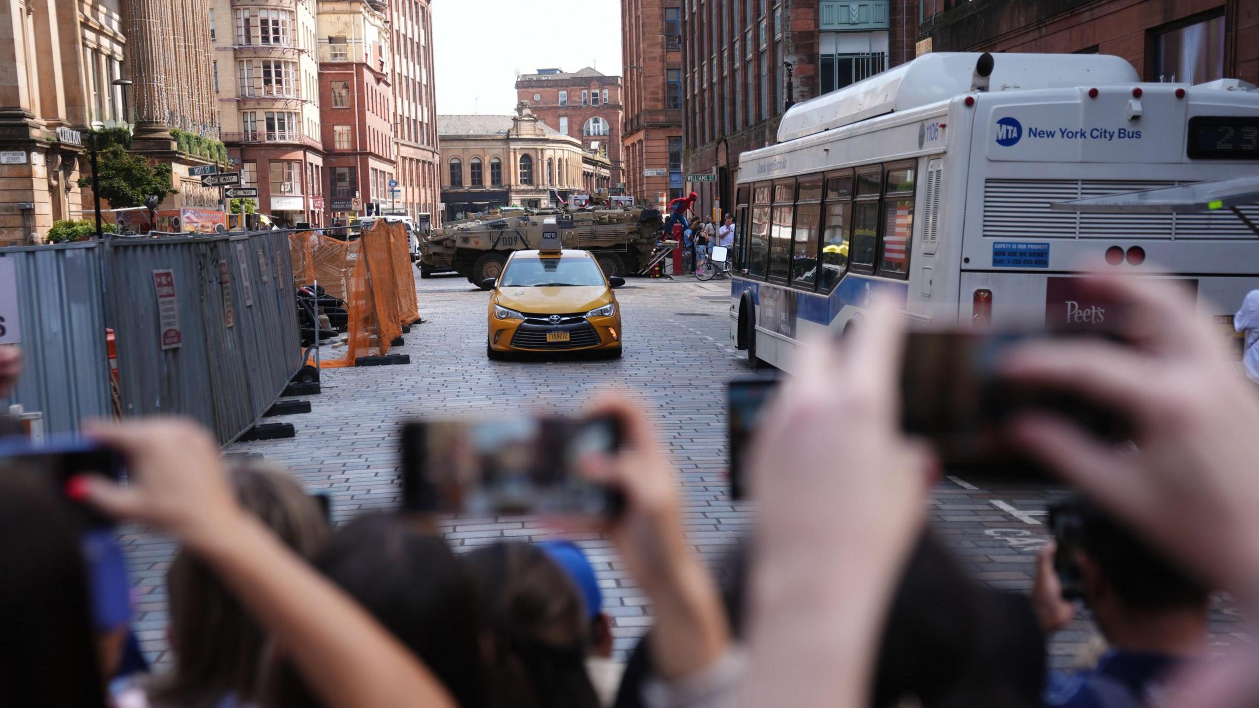 People take pictures as filming takes place in Glasgow. There is a white bus on the right and a yellow taxi in the centre of the image, with a tank in the background