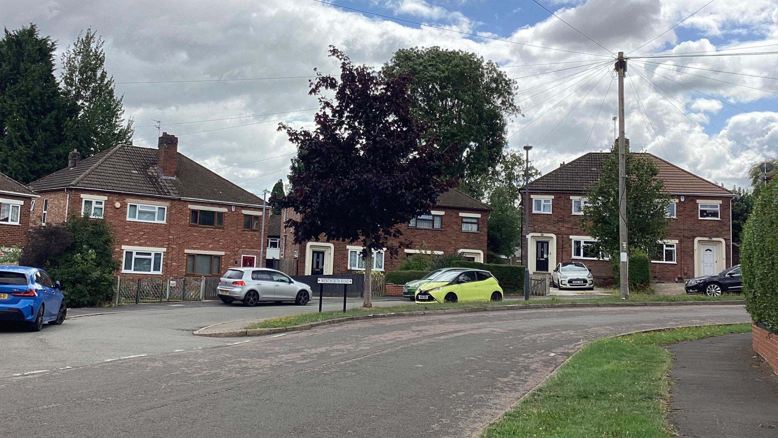 A road junction on a residential street lined with semi-detached houses. There are a number of cars parked on the road and on driveways.