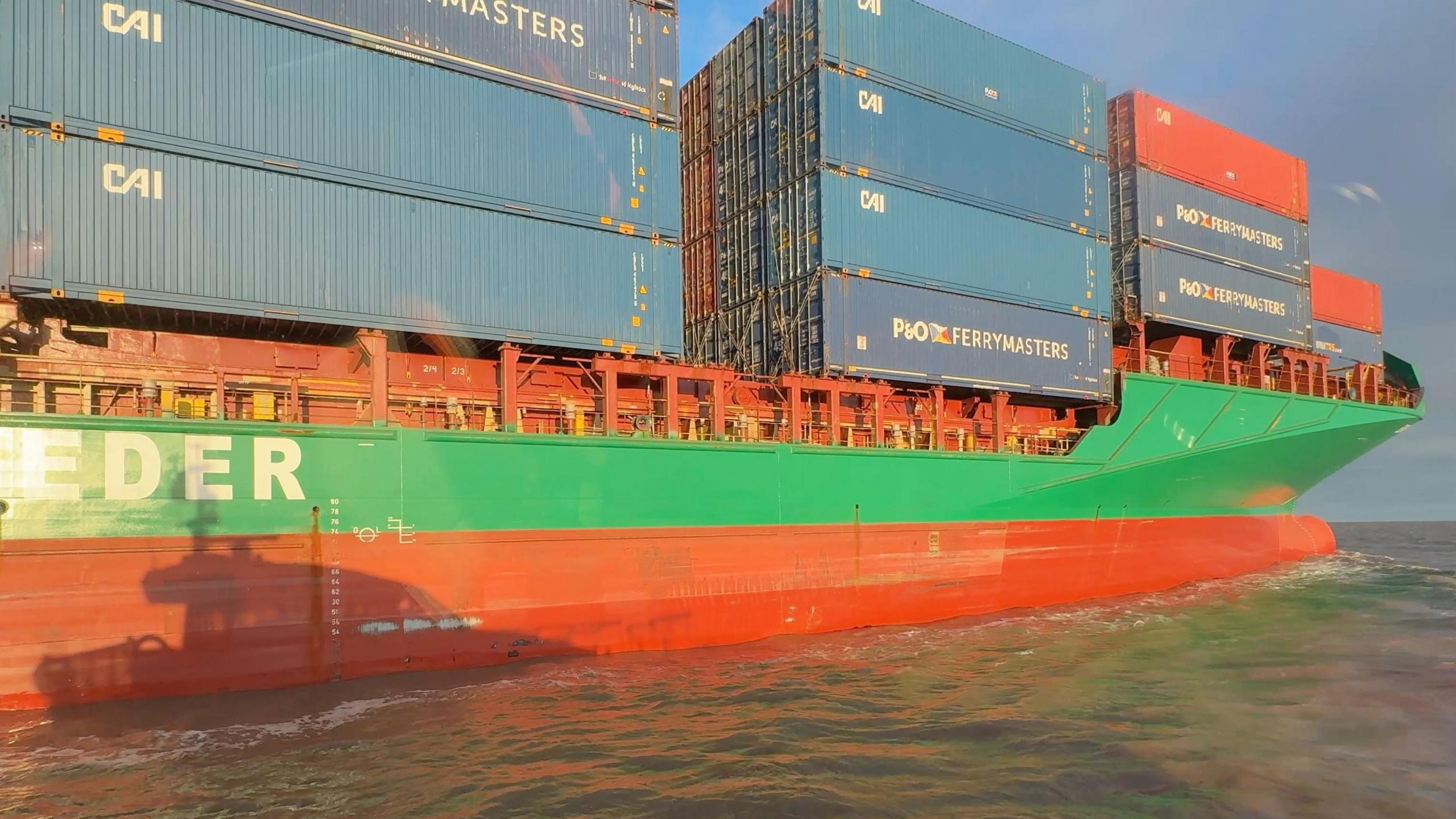 The shadow of a fast pilot launch can be seen against the red and green hull of a large cargo ship, which is carrying huge blue container boxes, stacked up to four high, on its deck.