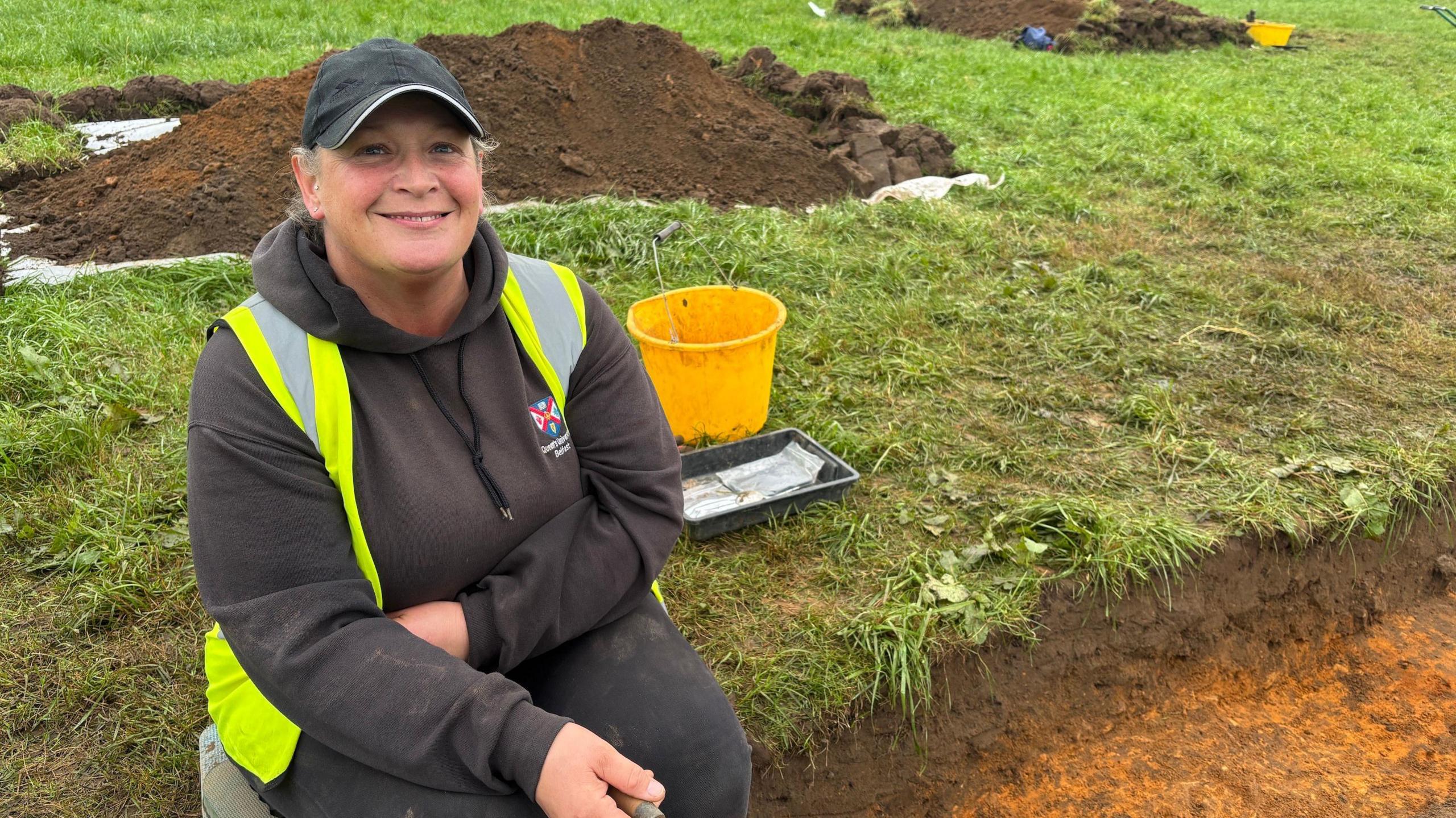 A woman sits in a field on the side of a dug out trench. She is dressed in black with a hi-vis vest on. behind her sits mounds of dug out soil and yellow buckets. She is smiling widely and holds a trowel in one hand. 