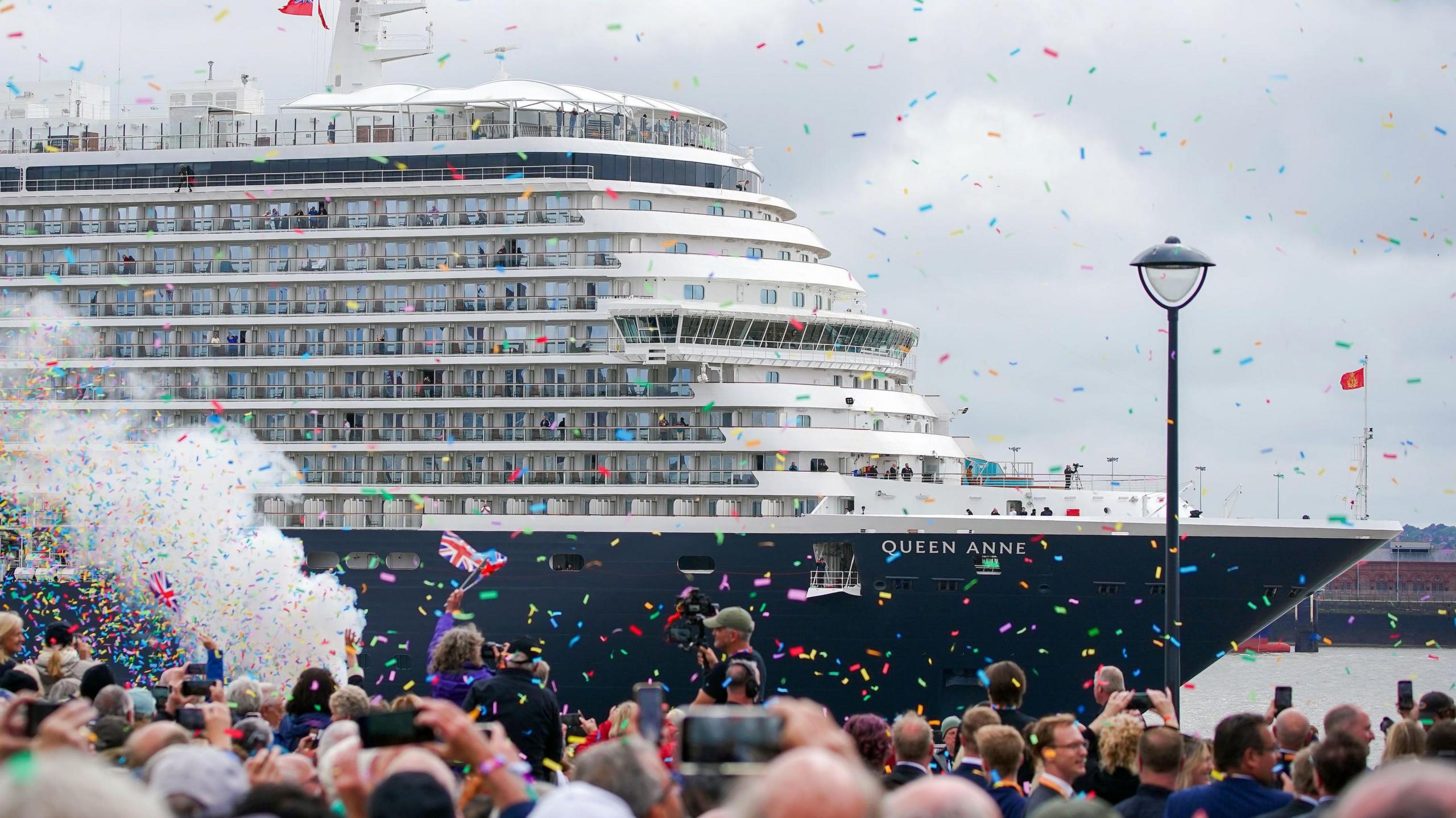 The many-storied Queen Anne cruise ship at a Liverpool dock surrounded by crowds at a celebration with multi-coloured confetti in the air 