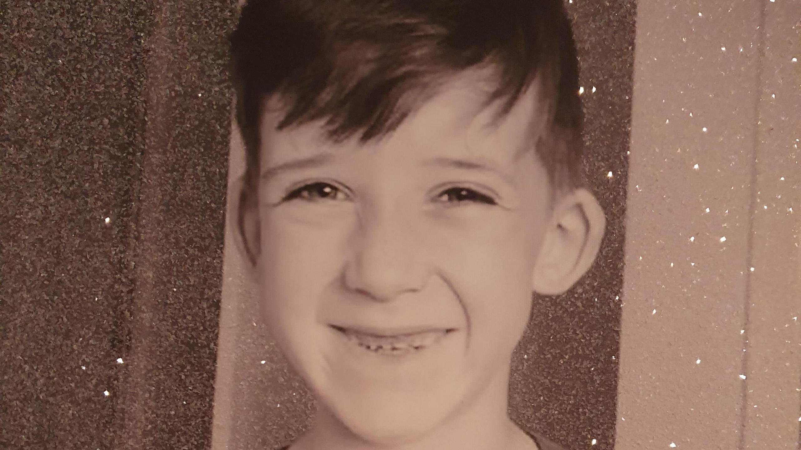 Kayden Walker looking directly at the camera. The picture is filtered with a brown-grey filter over the top and several white marks. Kayden is 12 years old and has dark eyes and dark hair covering his forehead.