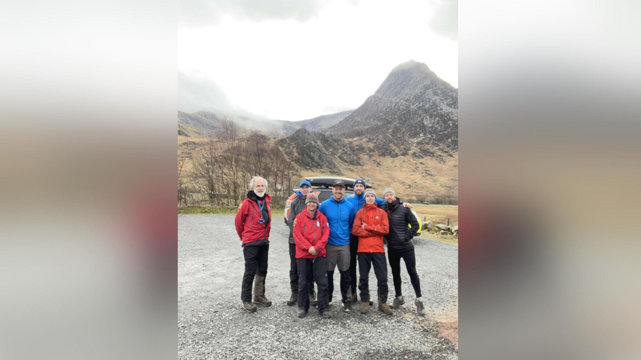 Mae grŵp o ddynion mewn siacedi coch, glas, oren a du yn sefyll gyda'u breichiau o amgylch ei gilydd o dan fynydd llwyd creigiog