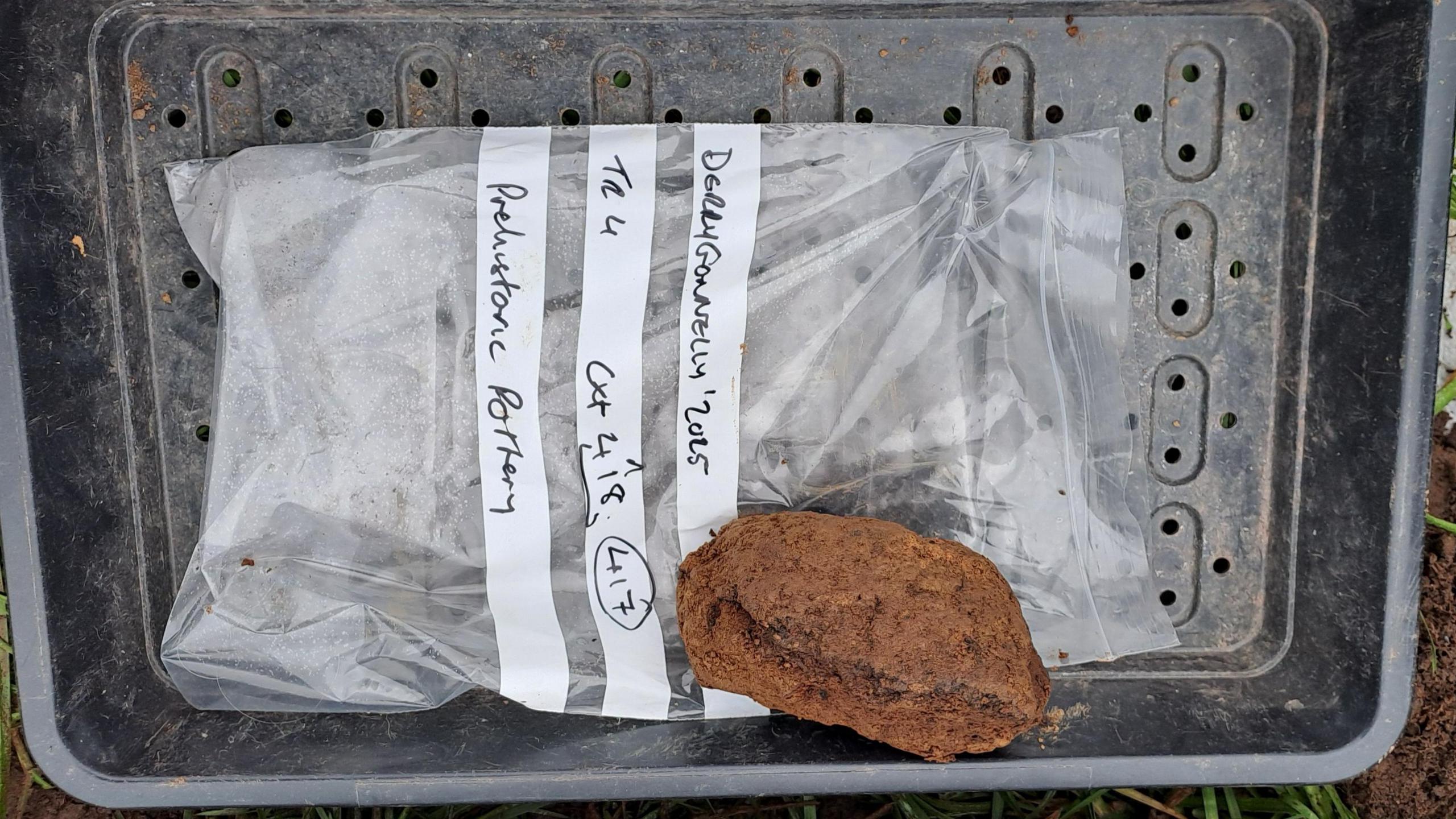 A black plastic tub with a clear plastic bag with information on it and a brown piece of pottery.