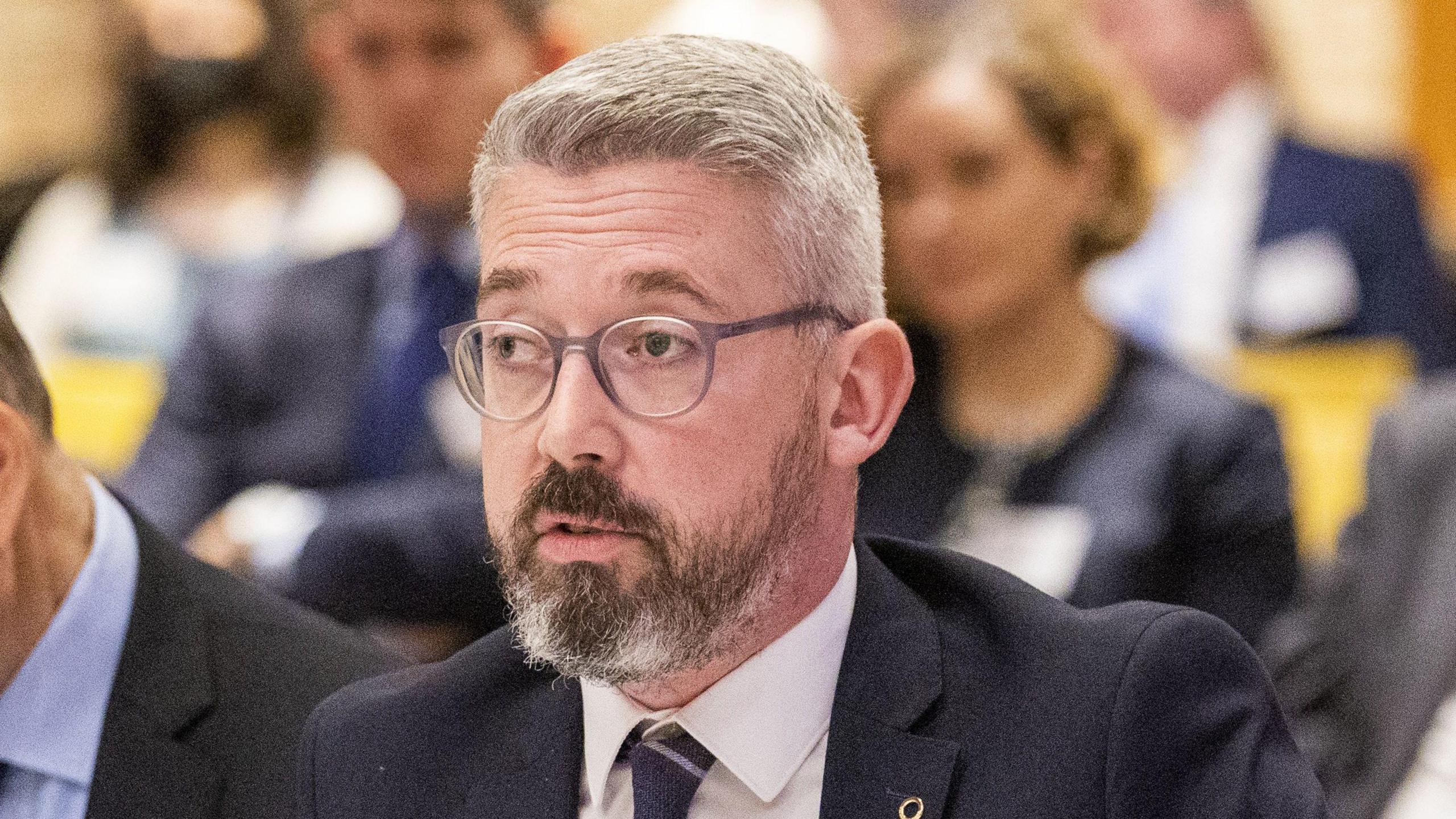 Niall Ó Donnghaile, picture from the chest up, wearing a navy suit, white shirt and navy tie. He is wearing glasses and has grey hair with a beard. 