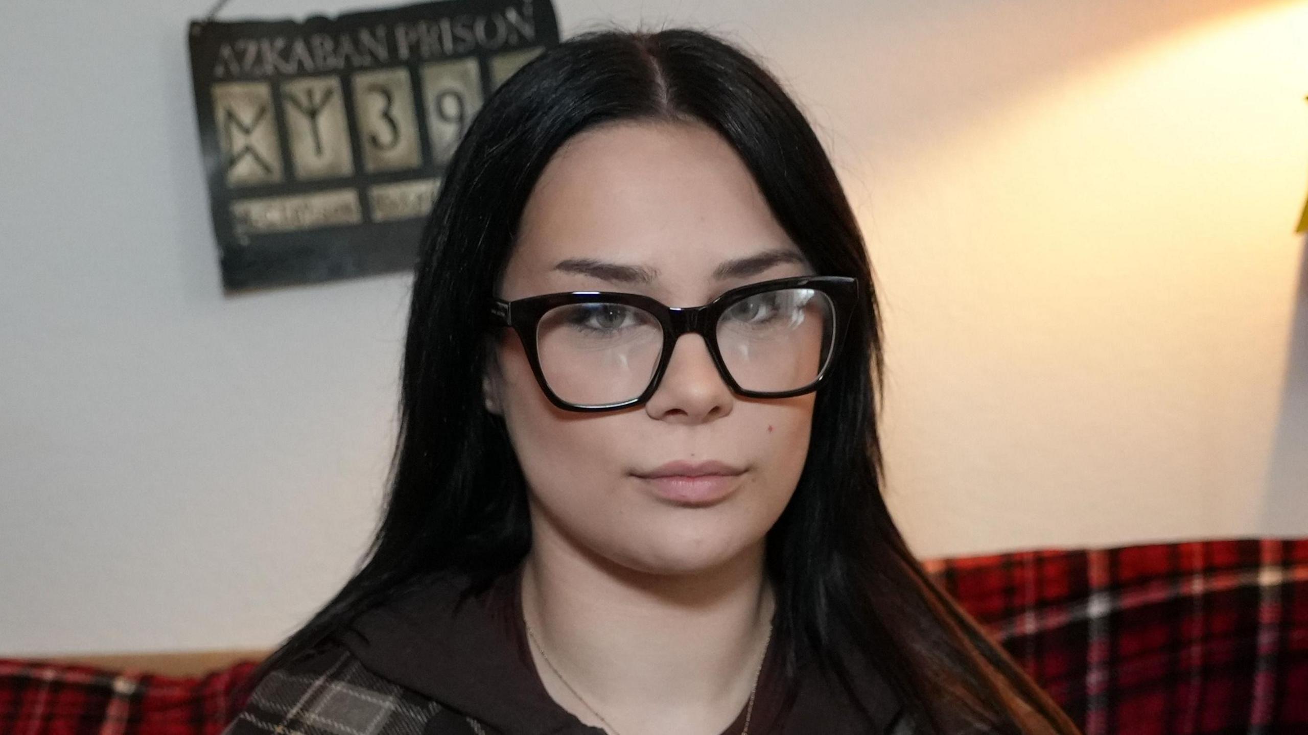 Poppy Stopforth slightly smiles at the camera. She has long dark hair, glasses and wears a grey and black chequered jacket. 