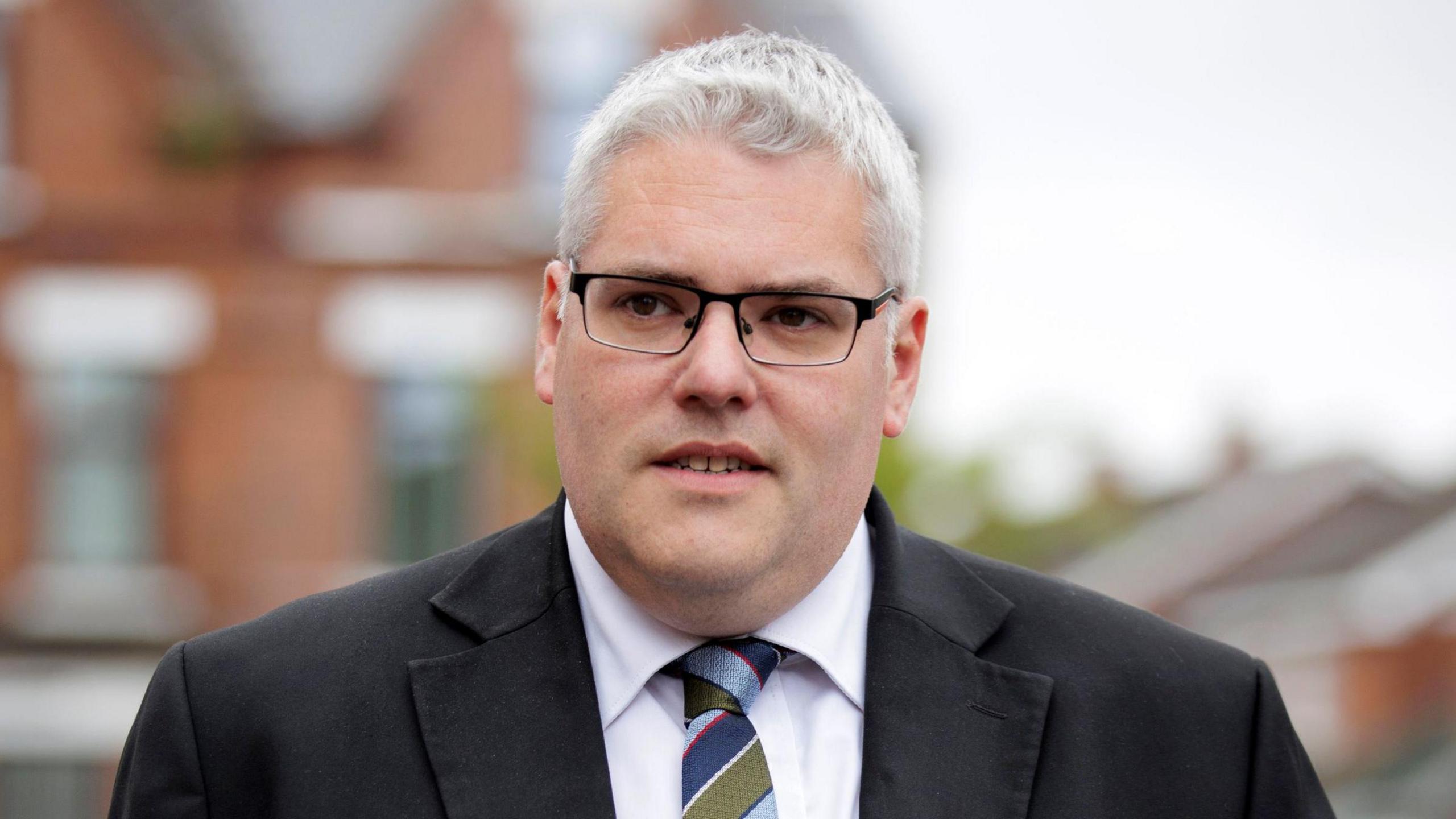 A man with short grey hair looks off to the side of the camera. He is wearing glasses, a dark suit jacket, white shirt and blue and green stripe tie. The background is blurred, but shows the outline of red brick buildings. 