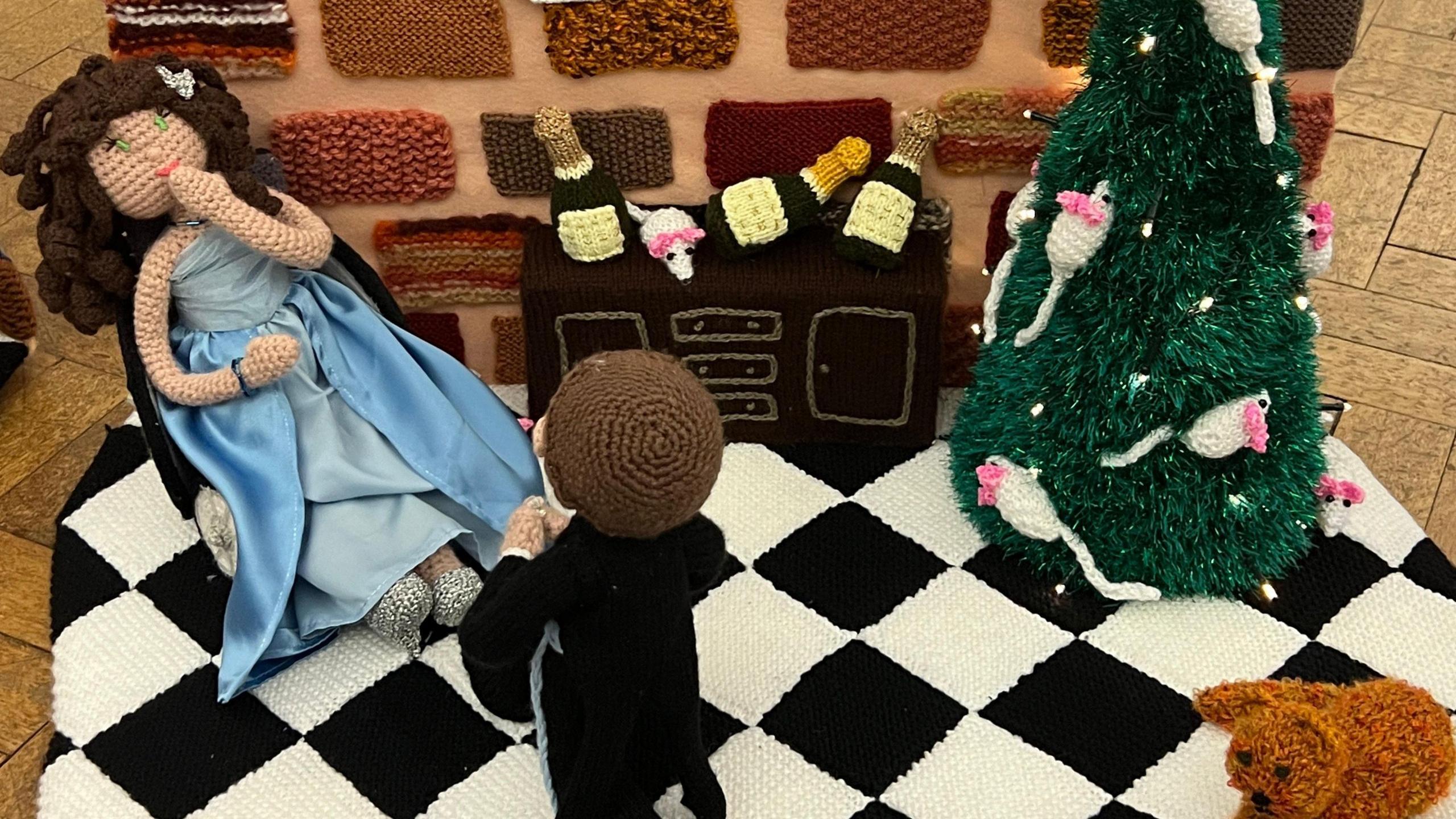 A woman in a knitted blue dress sitting down, with a man on his knees proposing to her. He is wearing a black tuxedo. There is a knitted ginger cat to the right and a Christmas tree with mice on it. 