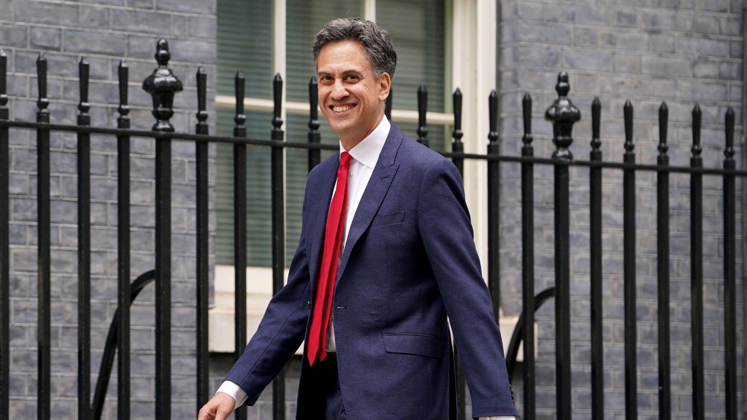 Ed Miliband smiling as he enters Downing Street. He has a blue suit and a red tie on.