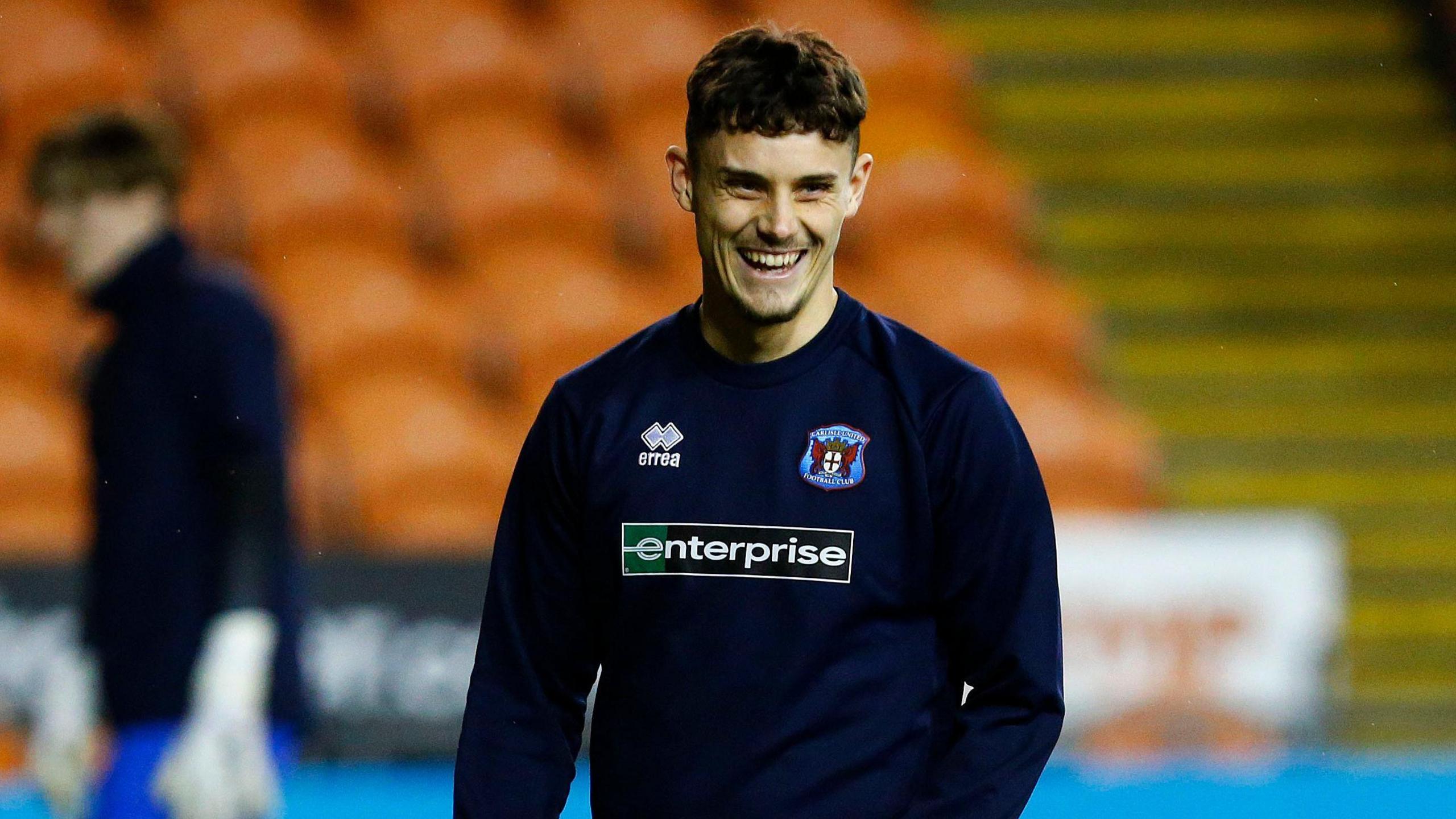 Cameron Harper warming up for a Carlisle game, wearing a navy blue club sweatshirt against a vague backdrop of orange seats