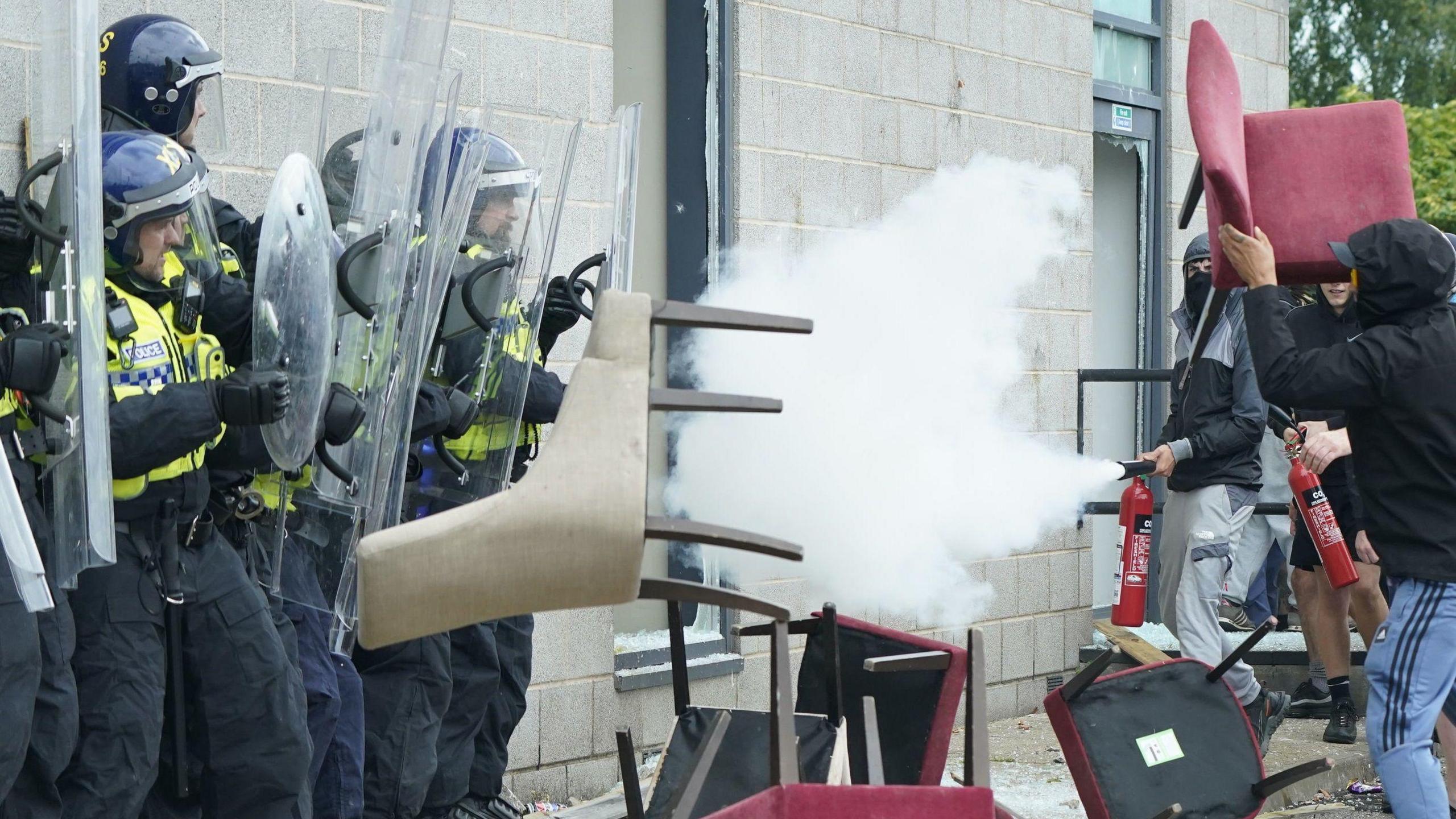 Protesters use fire extinguishers to attack officers. Chairs are thrown at the line of police officers who are holding up riot shields.