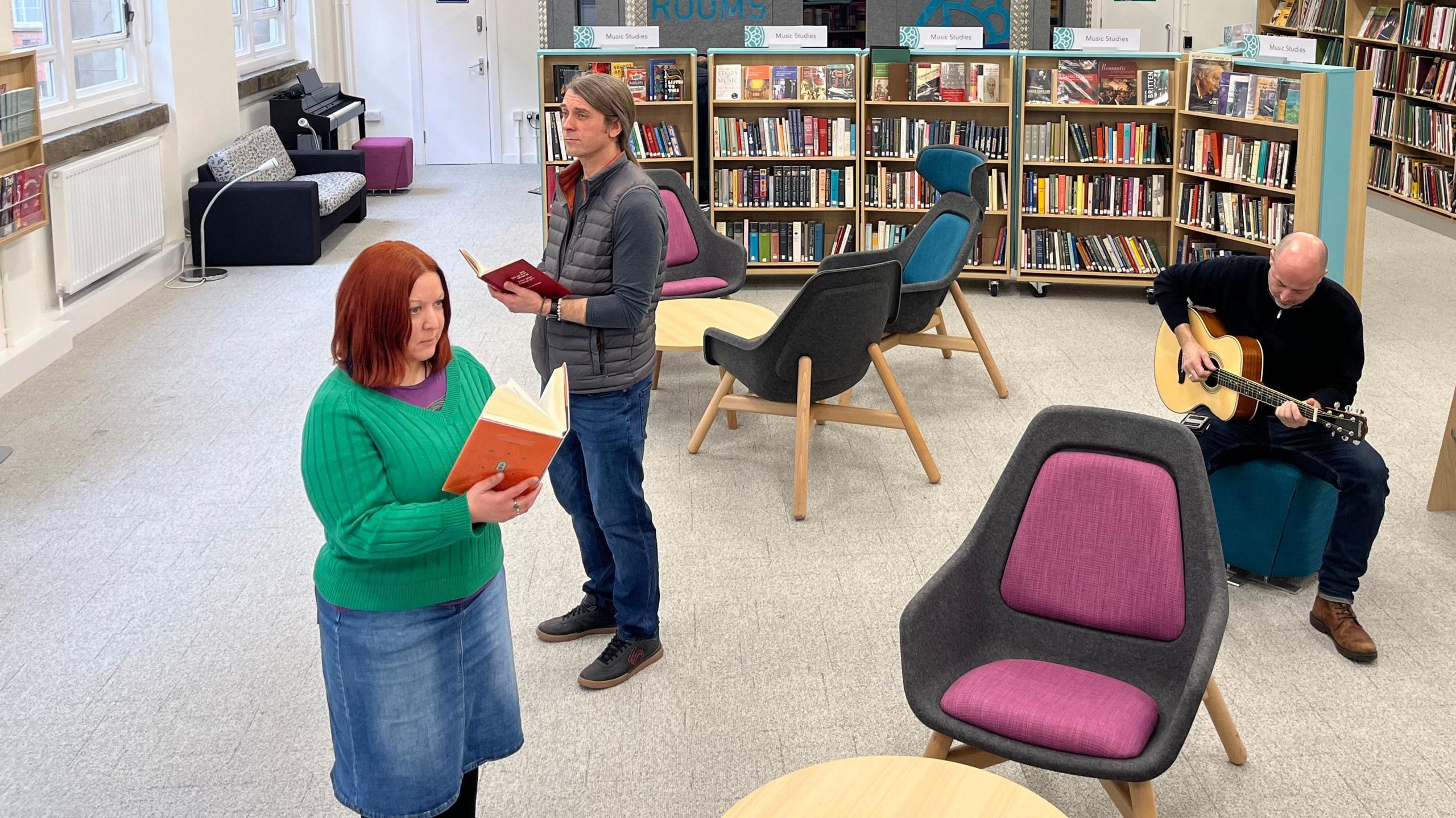 Three people, one playing guitar and two reading in a library