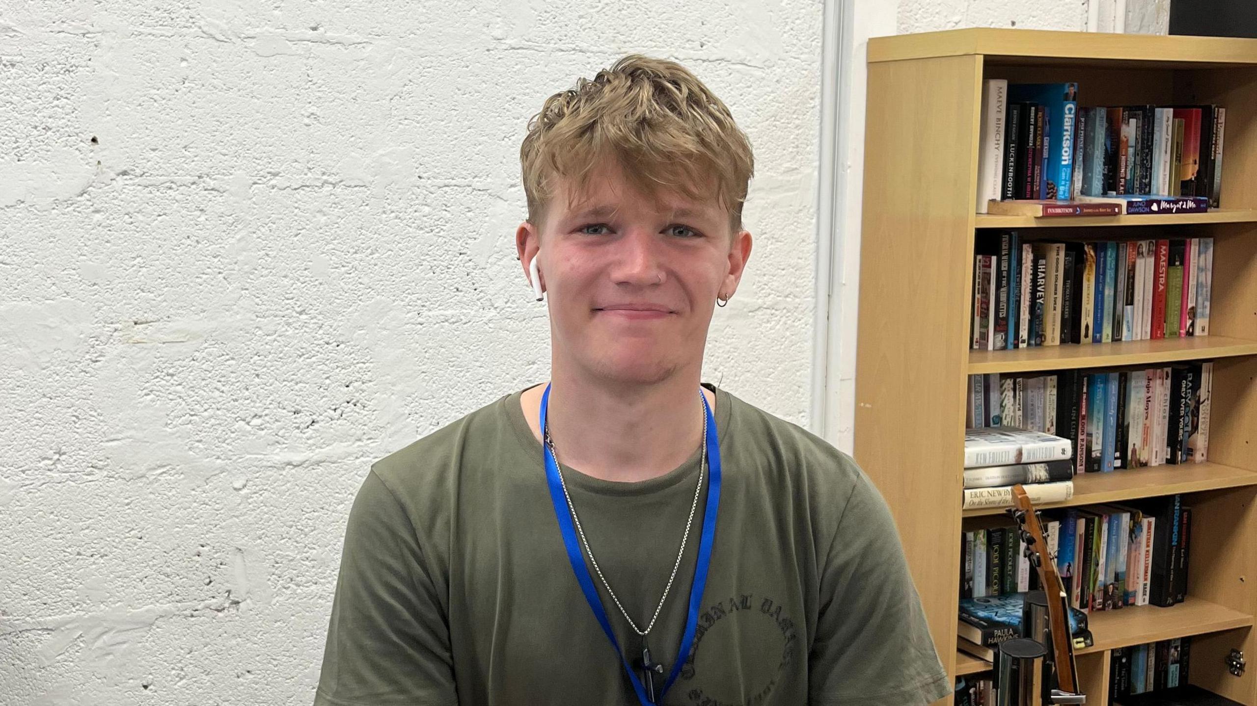 Volunteer Josh Lamphere smiles at the camera, he is wearing a khaki green t-shirt and a blue landyard and has an earphone in his right ear. Behind him is a book shelf full of books. 