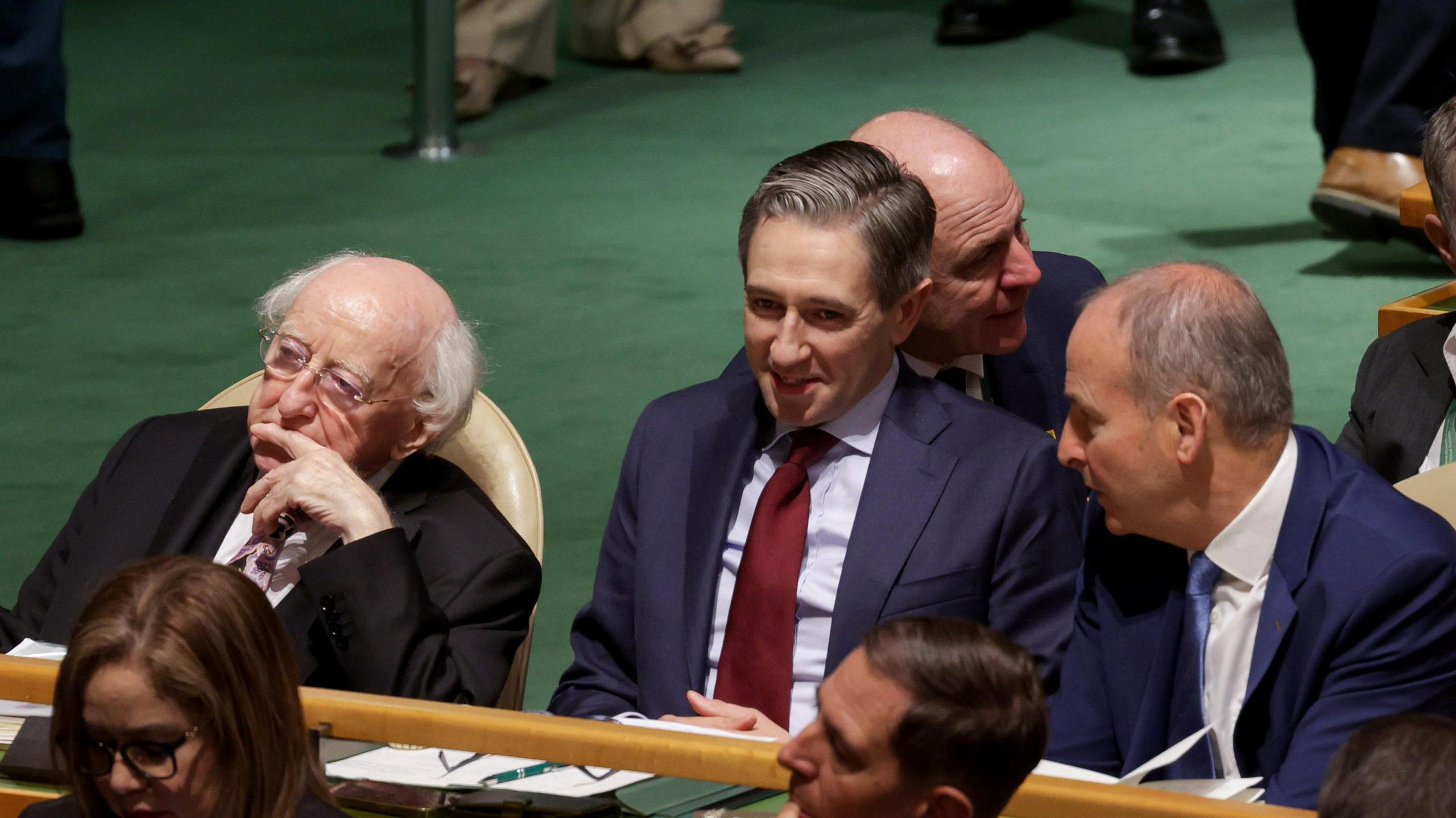 Three men, Michael D Higgins on the left wearing a black suit, white shirt and pink tie. In the middle is Taoiseach Simon Harris in a navy suit, blue shirt and blood red tie. In a navy suit, white shirt and blue tie on the right is Tánaiste Micheál Martin