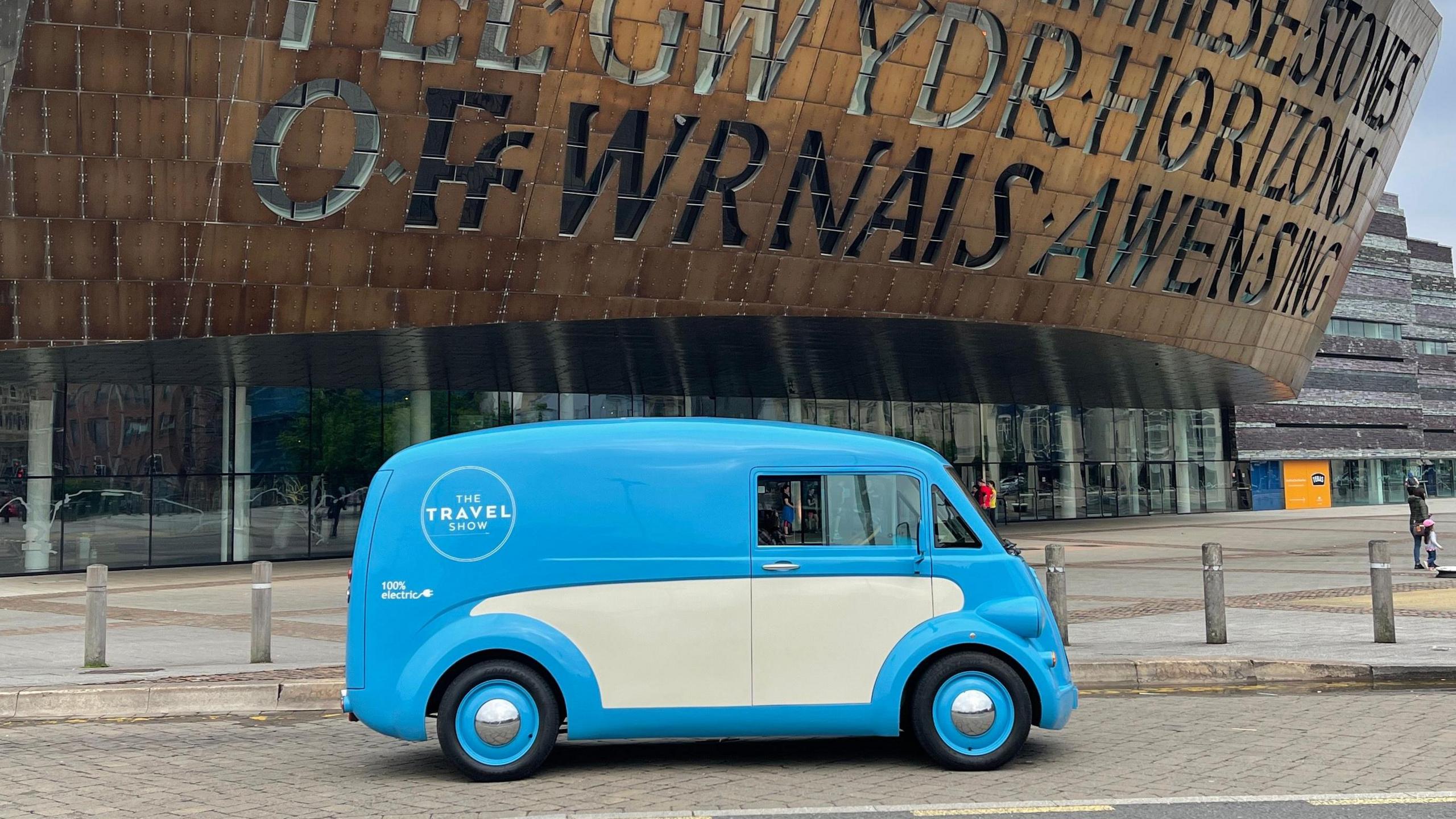 A retro looking blue and cream van with blue wheel trims is parked outside the Wales Millennium Centre. A logo on the side of the vehicles says 'The Travel Show'.