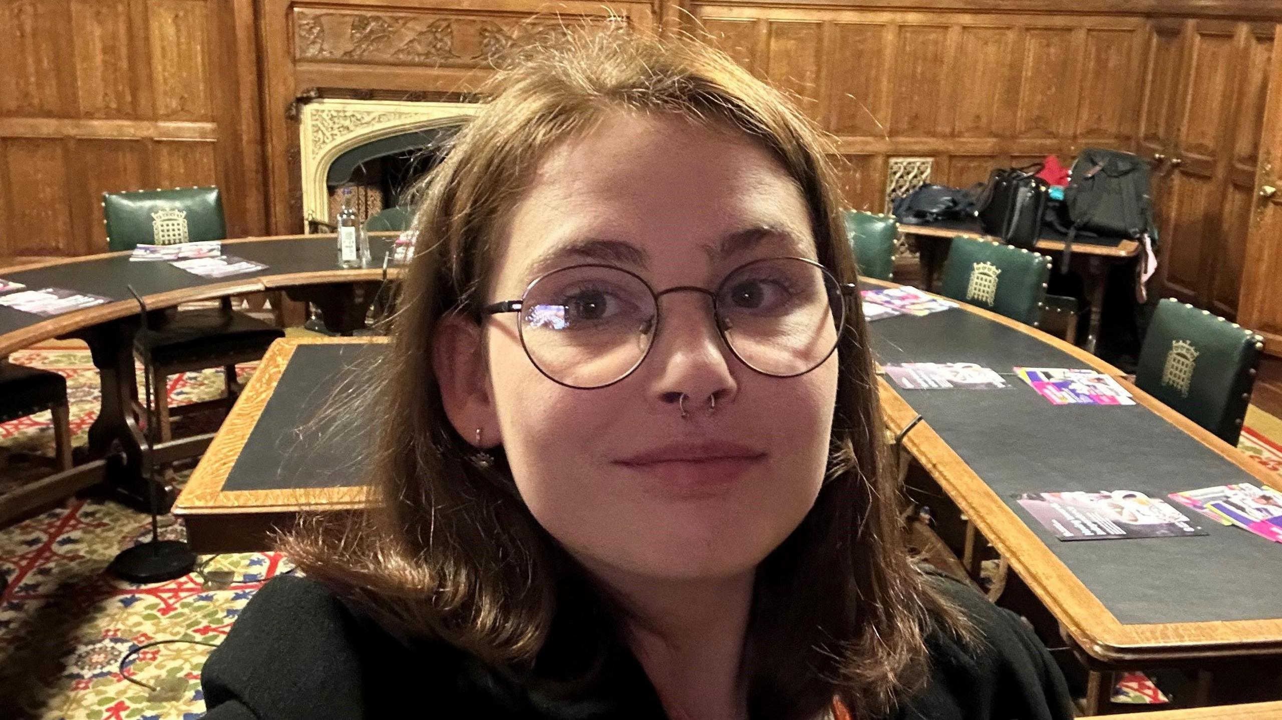 A young woman with brown hair and a septum piercing stands in a room in the House of Commons. Behind her are green leather chairs with gold crown emblems on them, and a long table.