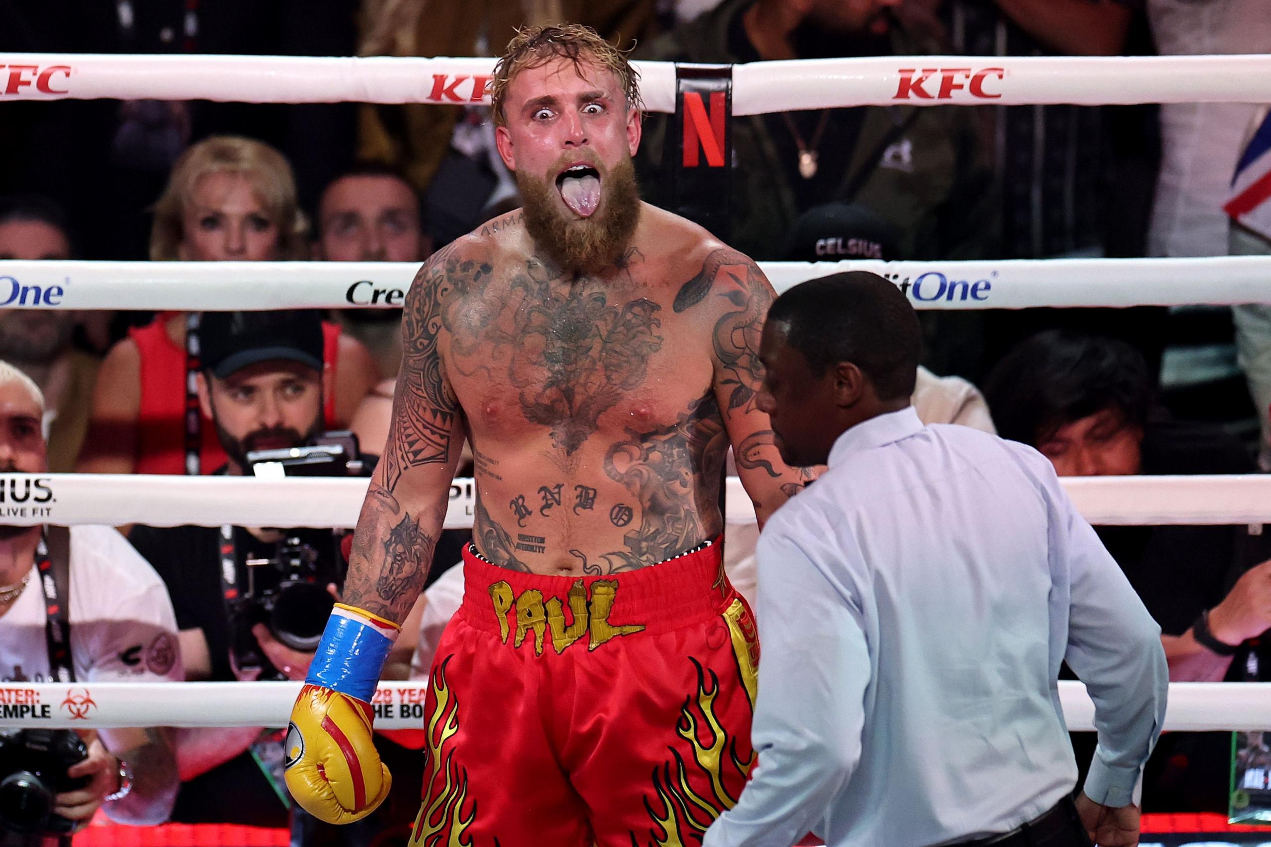 A tattooed boxer stands inside a boxing ring wearing bright red shorts with yellow flames and the name “PAUL” printed on the waistband. The boxer has yellow gloves and is shirtless, showing extensive tattoos on the chest and arms. A referee in a light blue shirt is positioned nearby, and several spectators and photographers are visible in the background outside the ring.