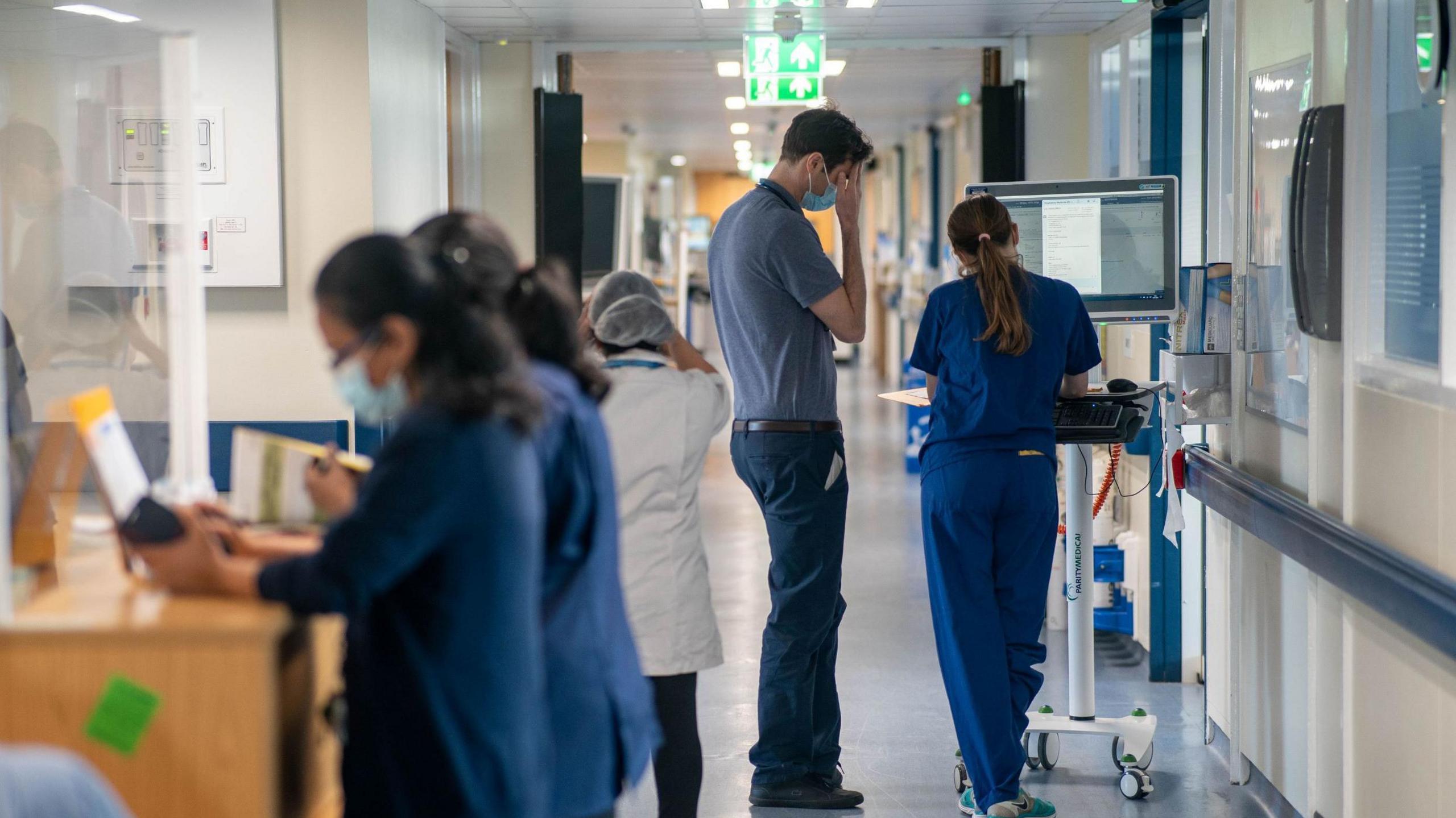 A generic picture of a hospital corridor, two female medical professionals in the foreground leaning on the desk of a nurses' station, an auxiliary walking away beyond them, and what looks like two doctors, a tall man and a shorter woman - in blue scrubs - leaning on a computer on wheels and looking at papers in a folder. 