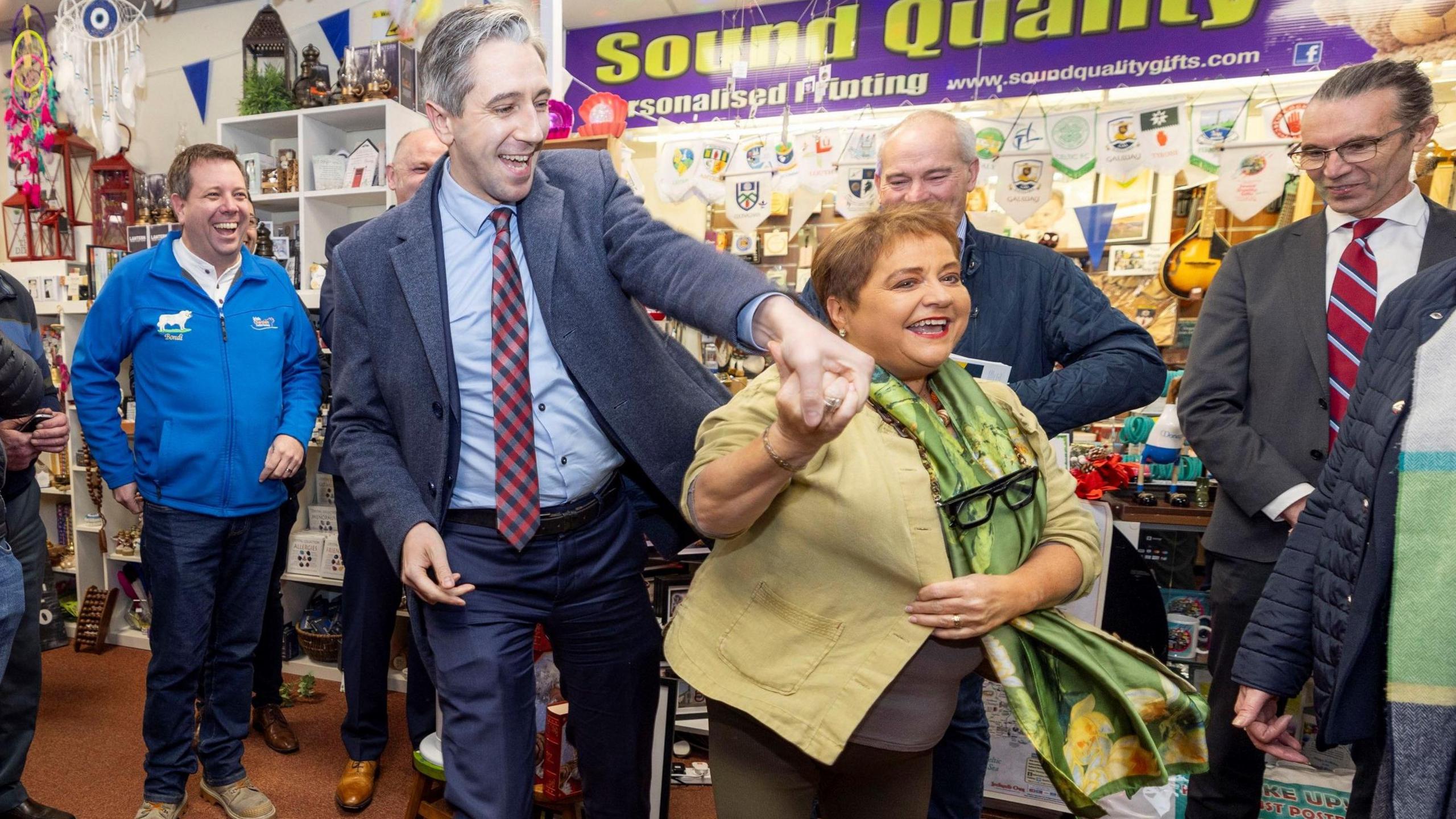 Taoiseach Simon Harris, wearing a navy suit, blue shirt and check red tie, smiling and holding hands with a shorter woman with short light brown hair. She is also similar and is wearing dark leggings, a cream shirt and a green silk scarf. They are surrounded by five smiling people. They are standing inside a gift shop. 