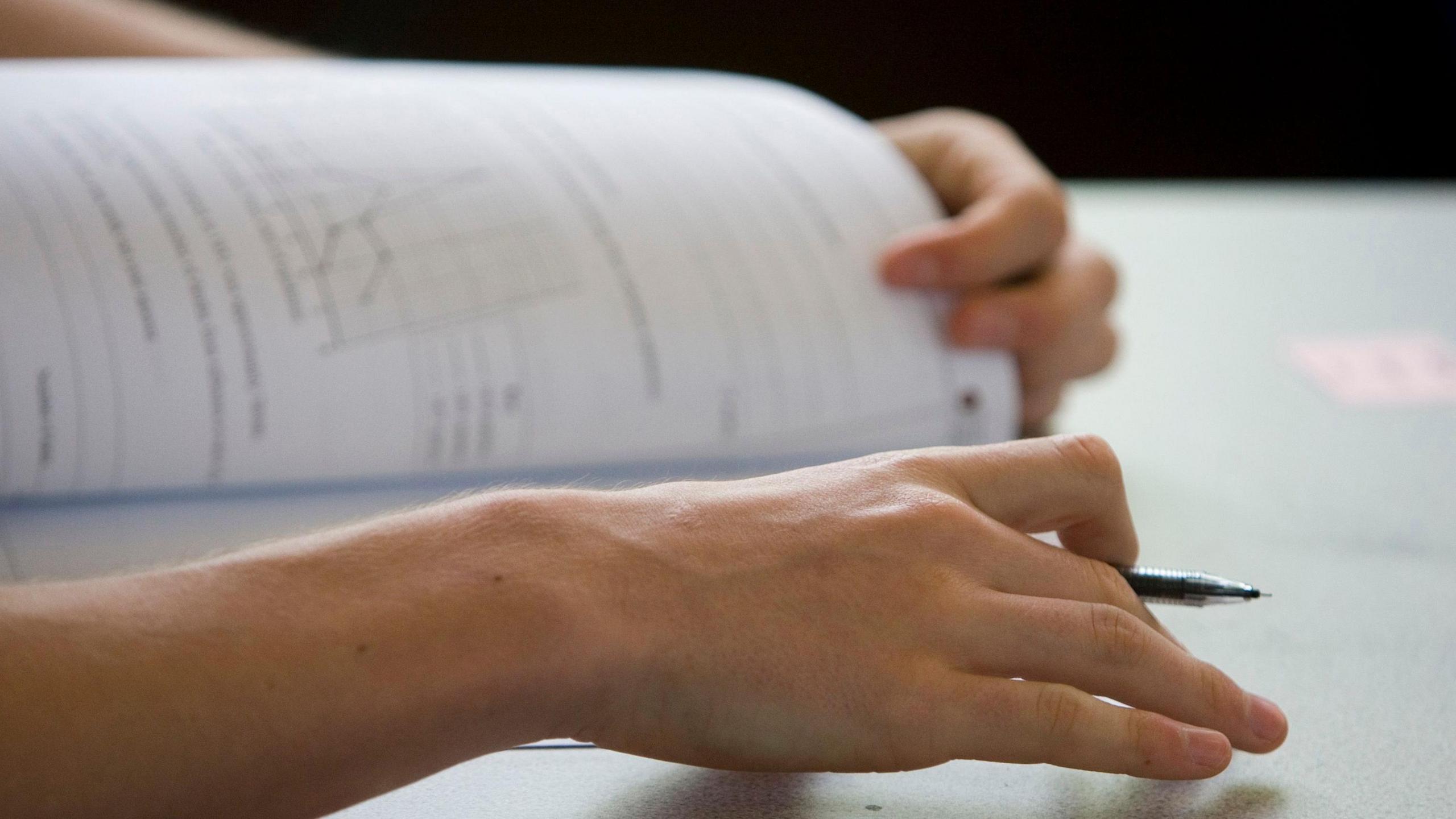 A student opens an exam paper with pen in hand. Only their hands are visible beyond the pen and paper.
