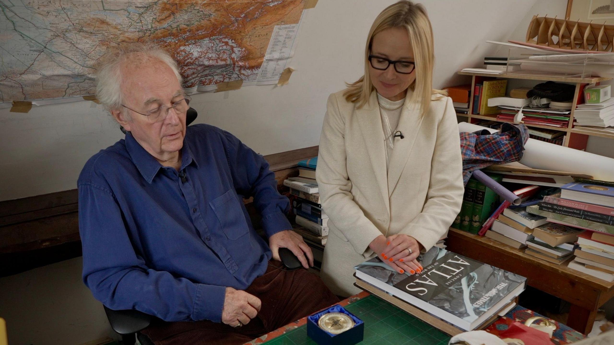 Philip Pullman and Katie Razzall in his study looking at his alethiometer, a mediums-sized circular gold object in a blue box lined with silk