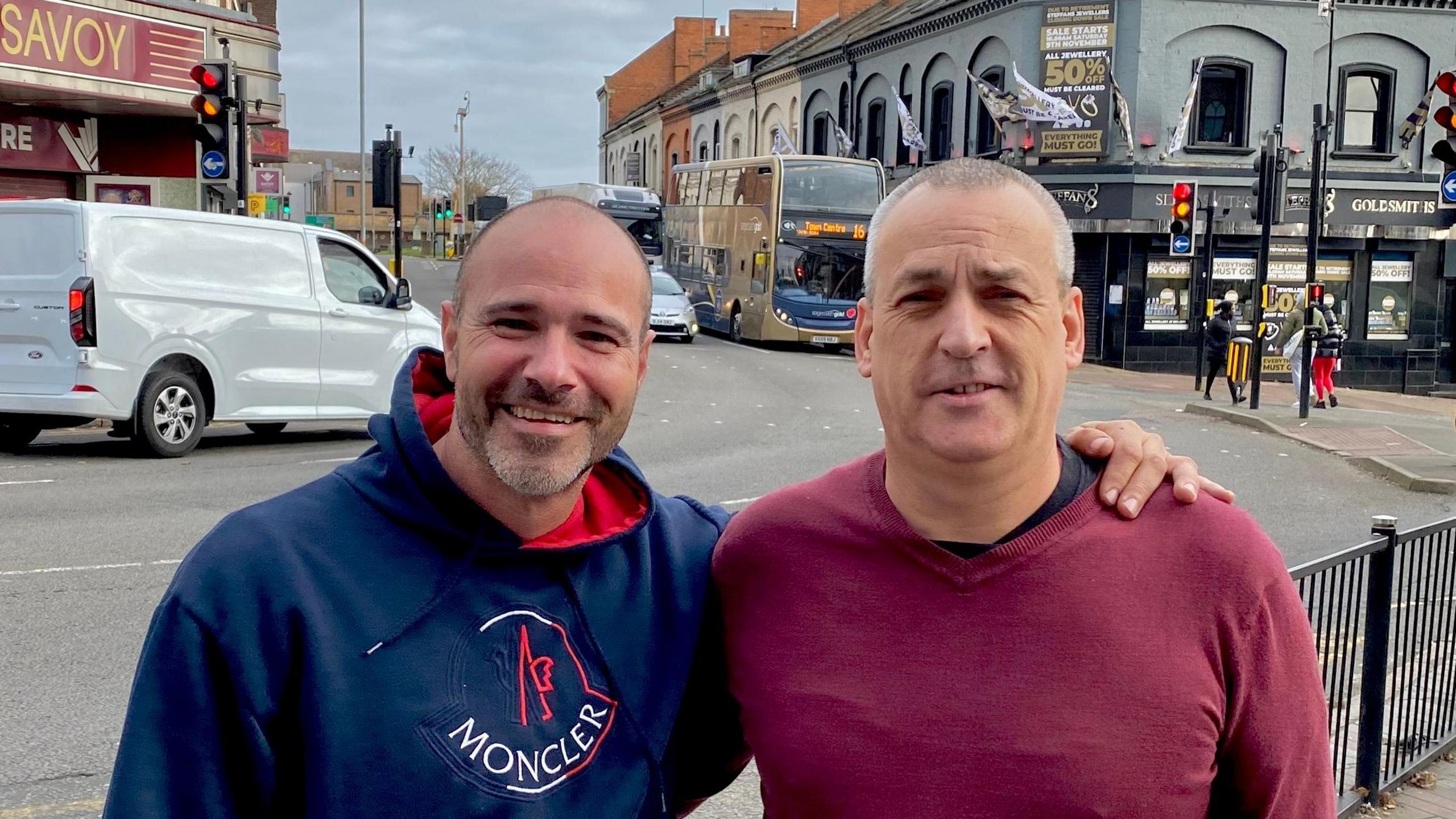 Justin Cohen in a blue hoodie, is stood with his arm round Steve McNeil, who is wearing a red top. They are standing on a pavement with a set of traffic lights and cars behind them. 