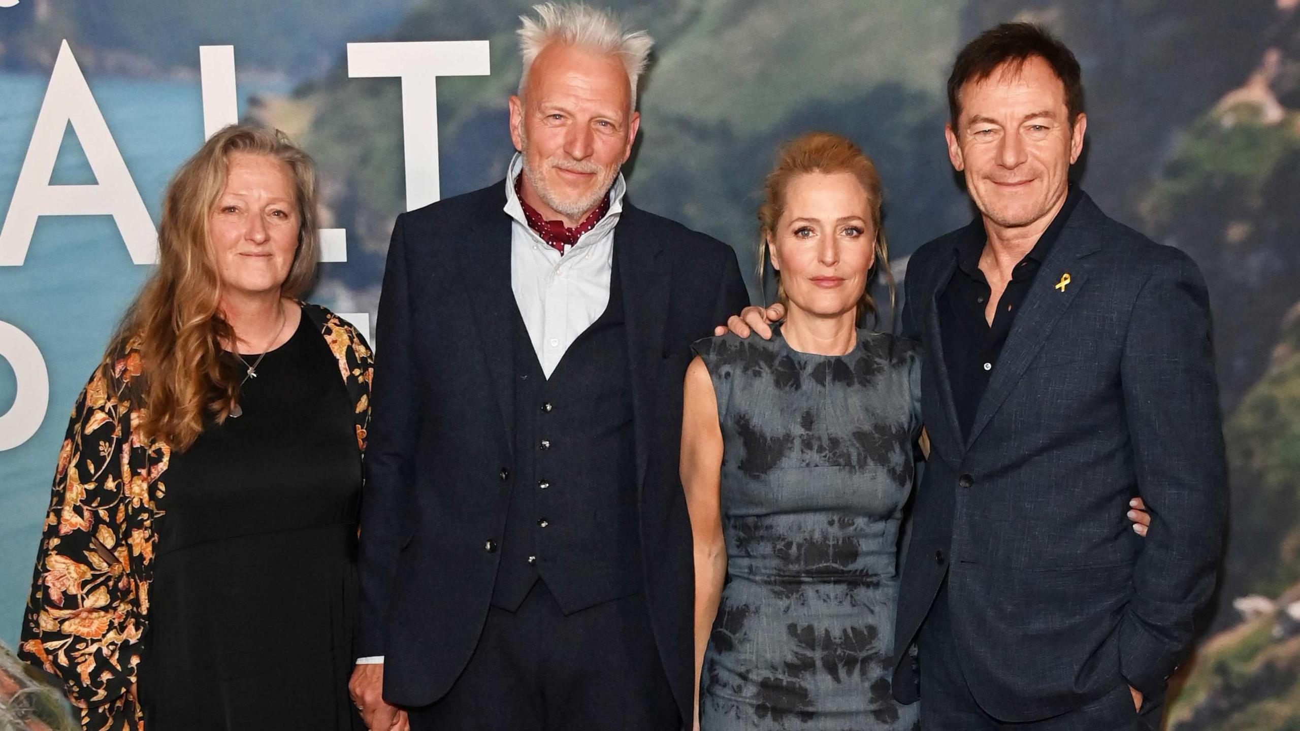 Raynor Winn, Moth Winn, Gillian Anderson and Jason Isaacs. All four of them are stood looking at the camera. Both middle-aged women are blonde, one male has white hair and the other has brown hair - both middle aged. Both men are in suites and the women are in dresses. 