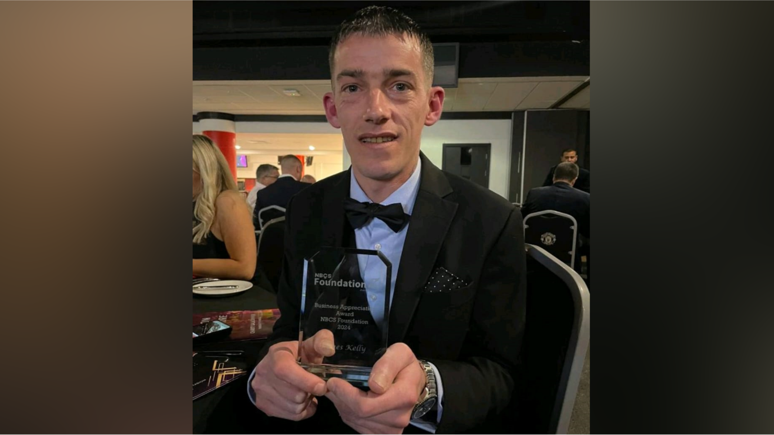 Image of James, in a suit and bow tie at an awards ceremony, holding up a glass trophy. 