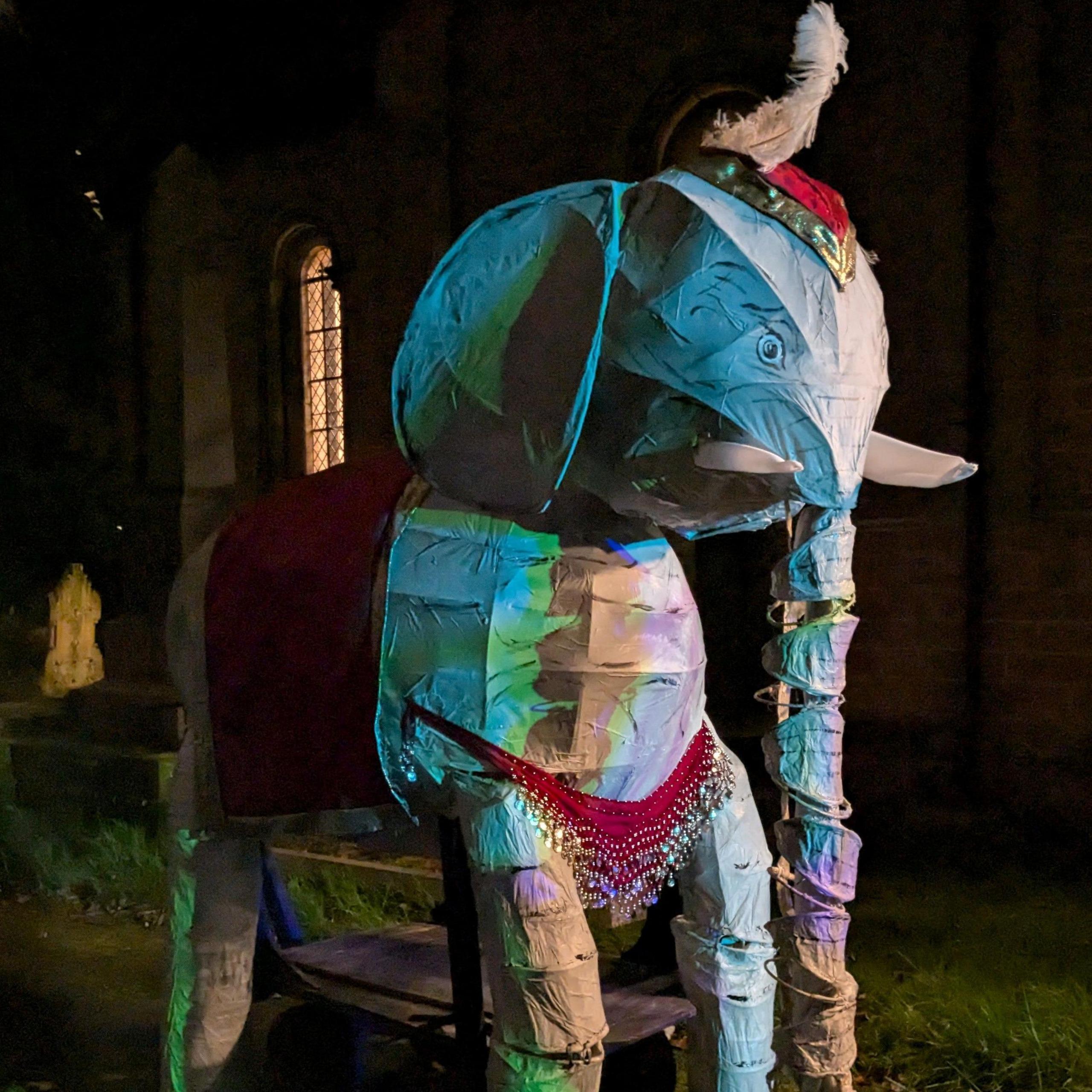 A wire frame model of an elephant, covered in multi-coloured fabrics and topped by an Indian-style head covering and a feather sticking out.