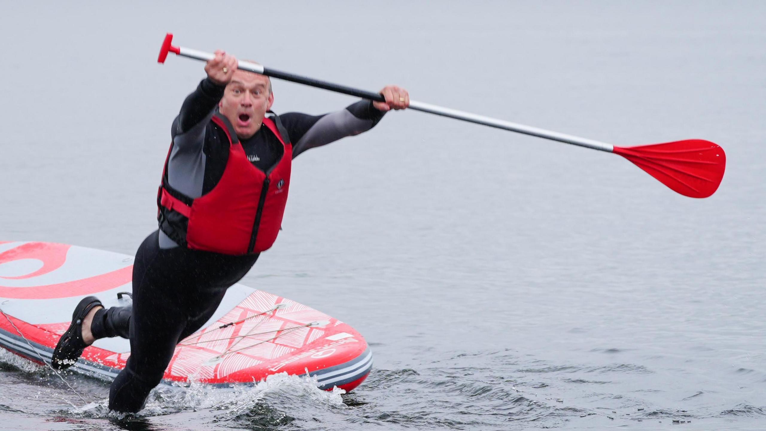 Ed Davey fakes surprise as he falls into the water from a paddleboard, wearing a black wetsuit and red life preserved and holding a paddle