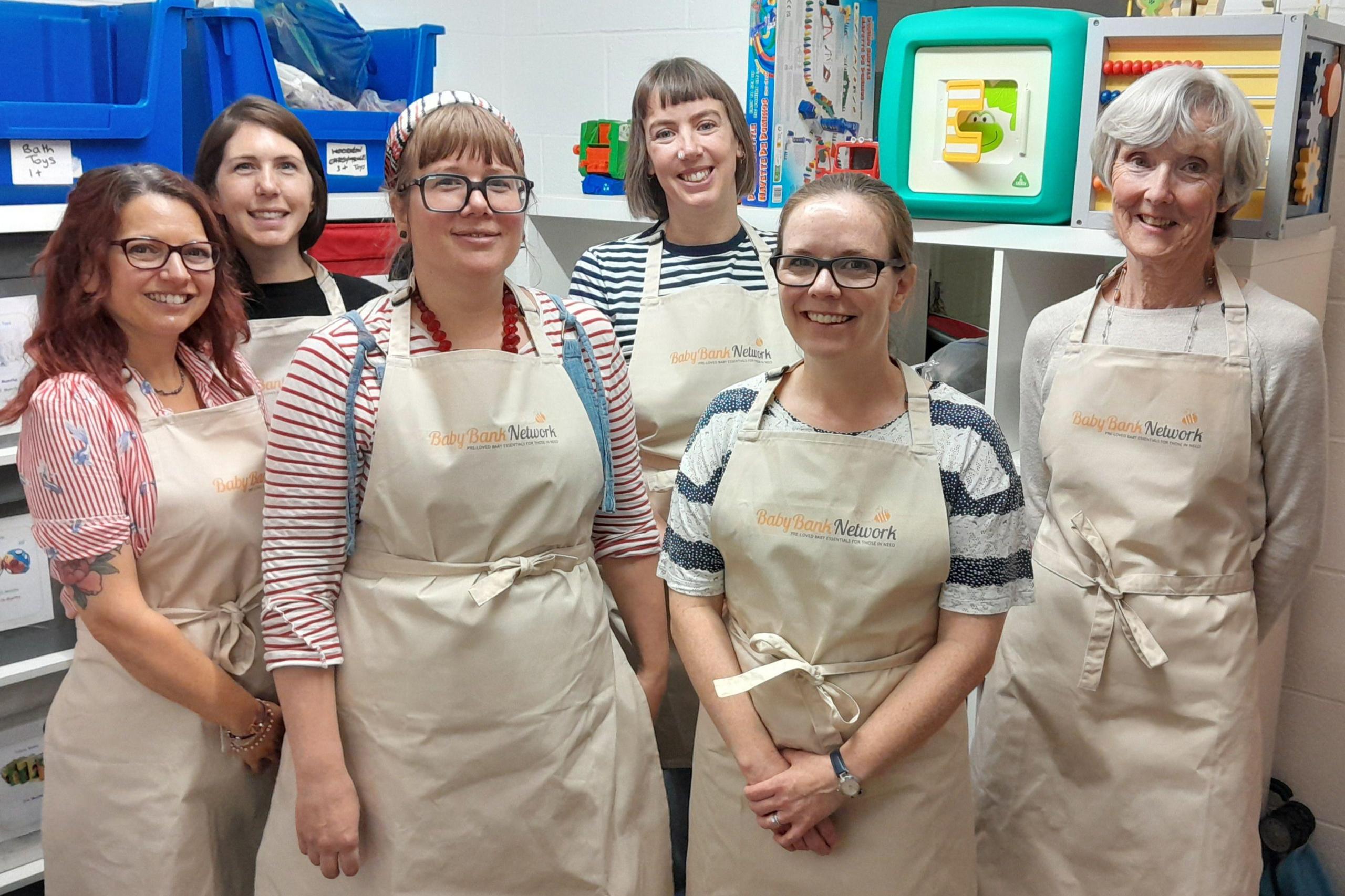 Image of six women in a toy storage room. They are dressed in tan aprons tied in bows at the front with the words "Baby Bank Network" on the front of each apron.