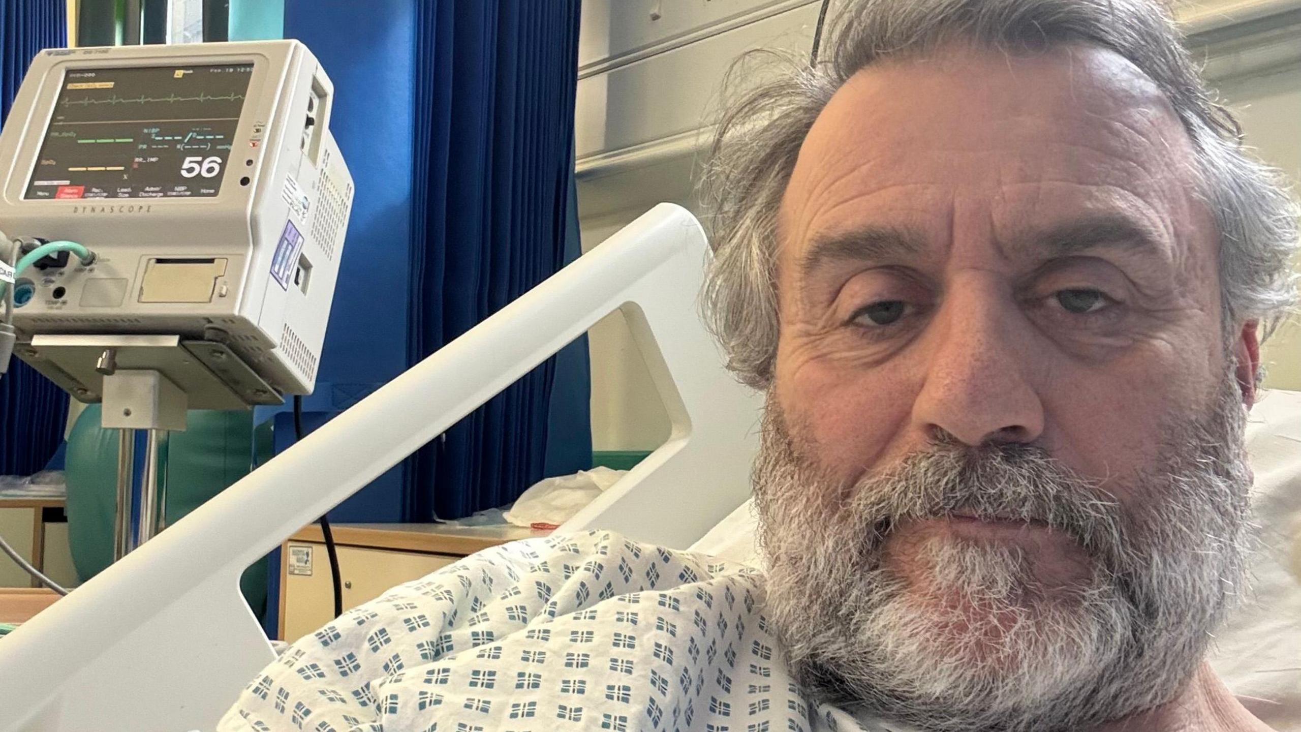 Robert with a full grey beard and grey hair, in a hospital bed, with a machine next to him, looking at the camera and wearing a hospital gown. 