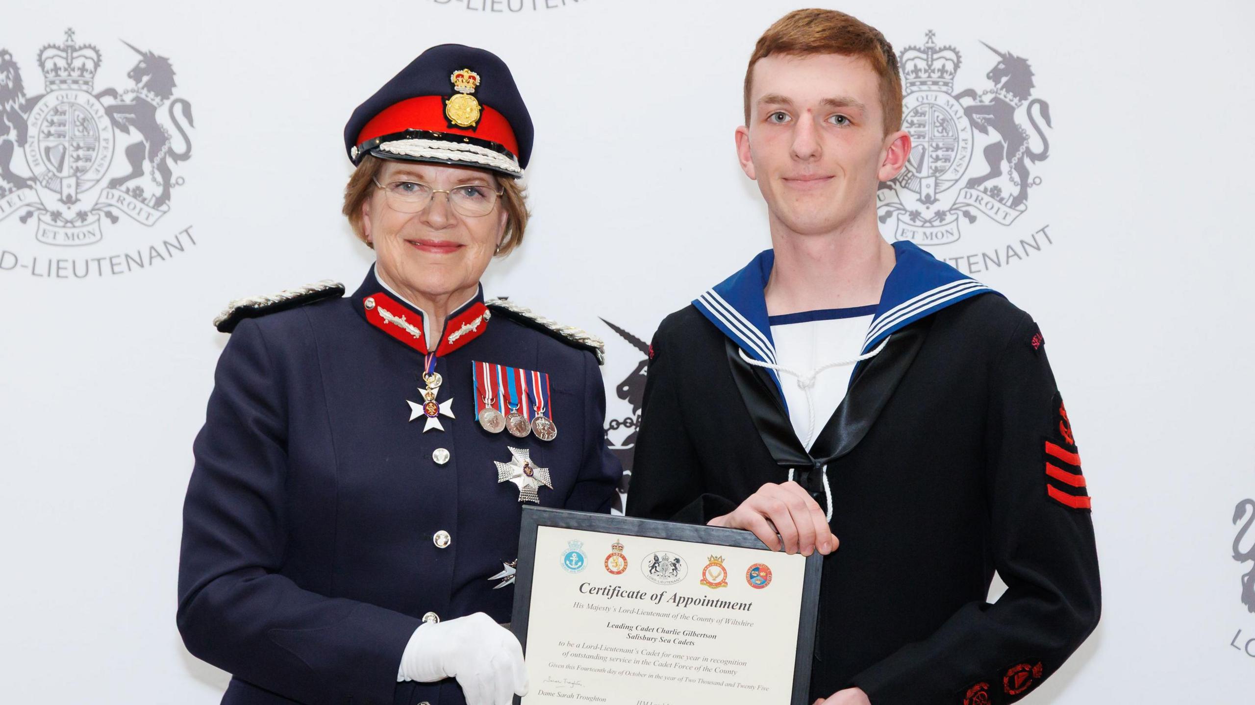 Dame Sarah Troughton wearing military jacket and medals with Charlie in a black jacket and holding a certificate.
