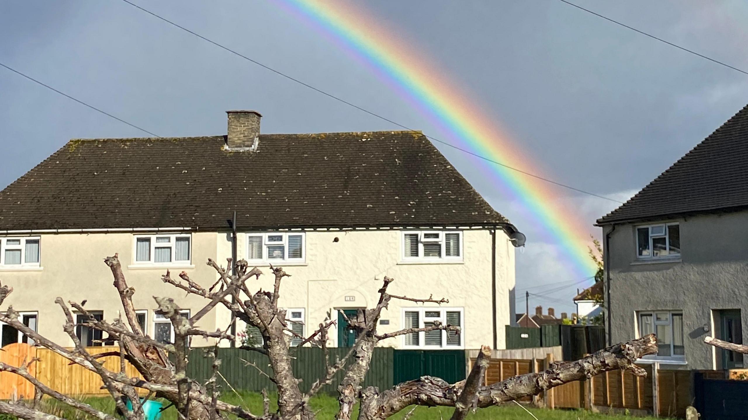 Two sets of semi-detached houses with cream render dominate this picture. A bright strand of a rainbow fills the gap between the buildings with dark skies behind it. A heavily pruned tree is in the foreground.