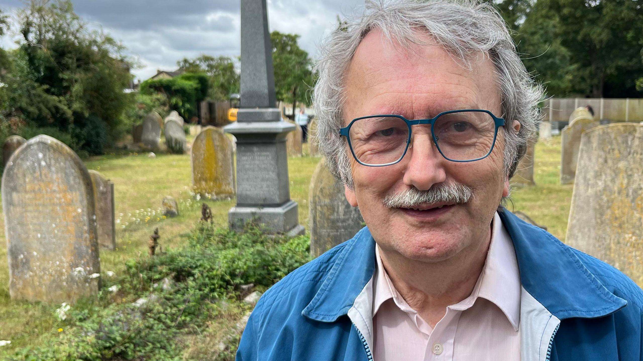 David Edwards is standing in the cemetery and there a gravestones on the grass behind him. He has medium length grey hair and is wearing blue framed glasses that match the colour of his blue jacket. He has a moustache and is also wearing a pink and white shirt.
