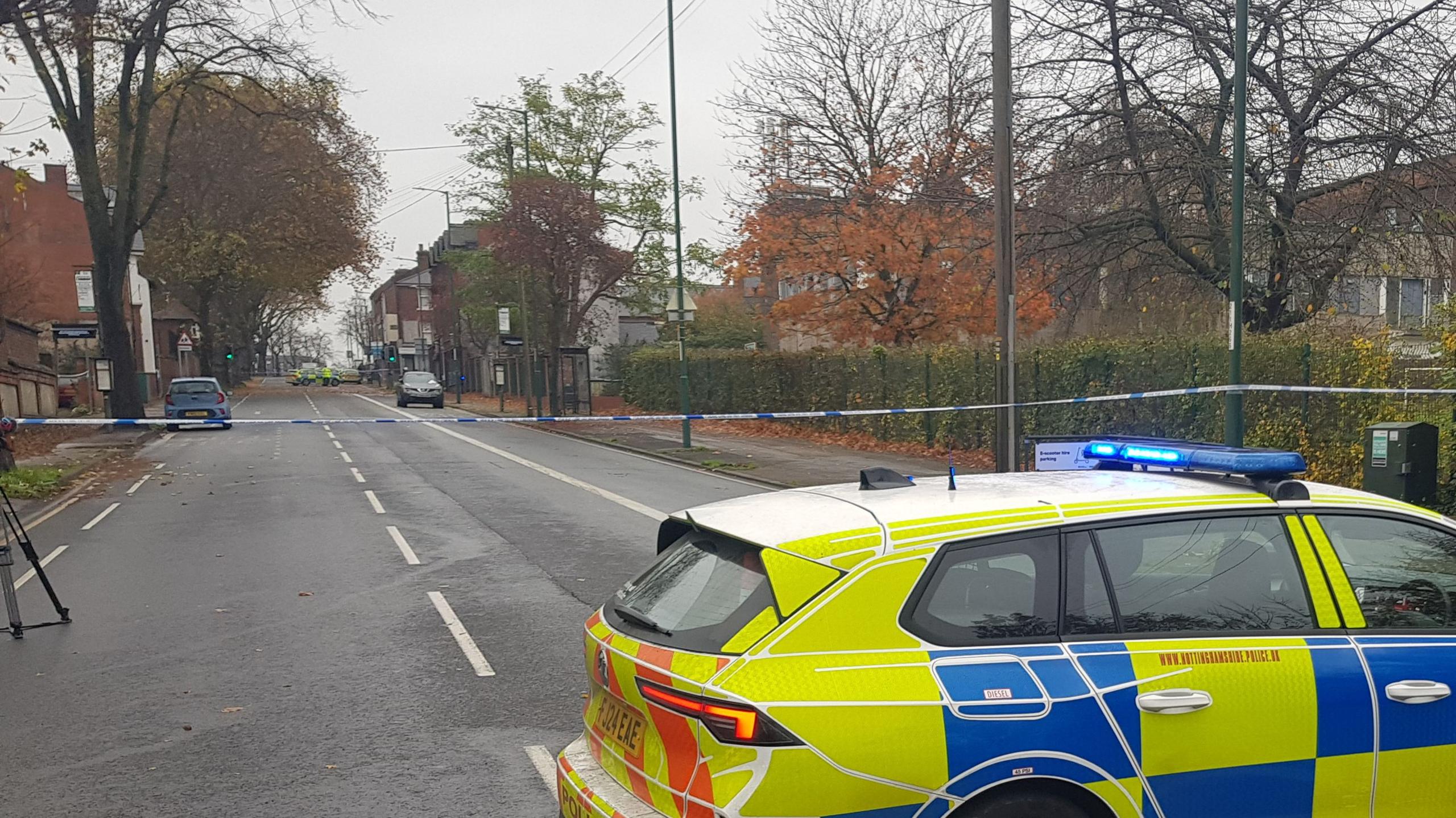 A police car is parked in front of a police cordon across the road. Police vehicles and staff can be seen within the cordon.